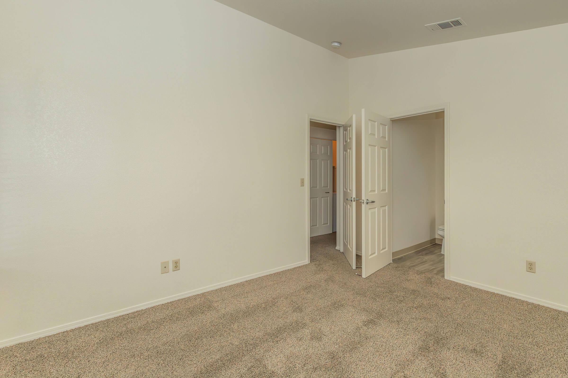 A spacious, empty room with beige walls and carpeted flooring. There are two doors visible: one leads to another room and the other may lead to a bathroom. Natural light illuminates the space, highlighting the clean and simple design.