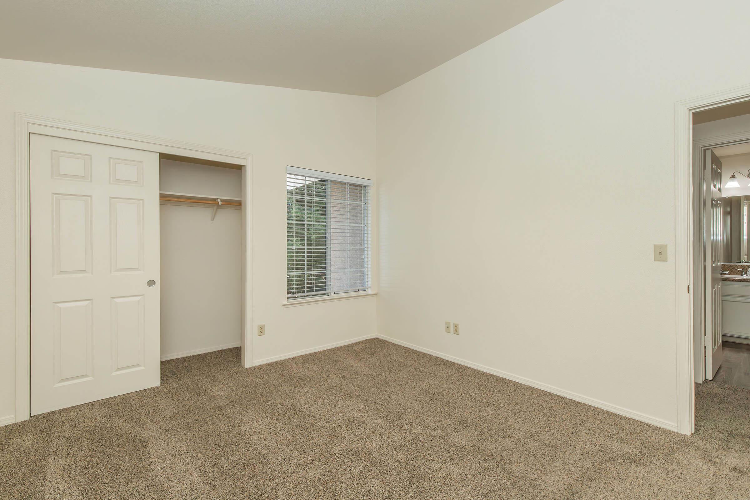 A vacant room with beige carpet, white walls, and a window with blinds. There is a closet with double doors on the left side and a door leading to another area on the right. The room has no furniture and is well-lit, creating a spacious feel.