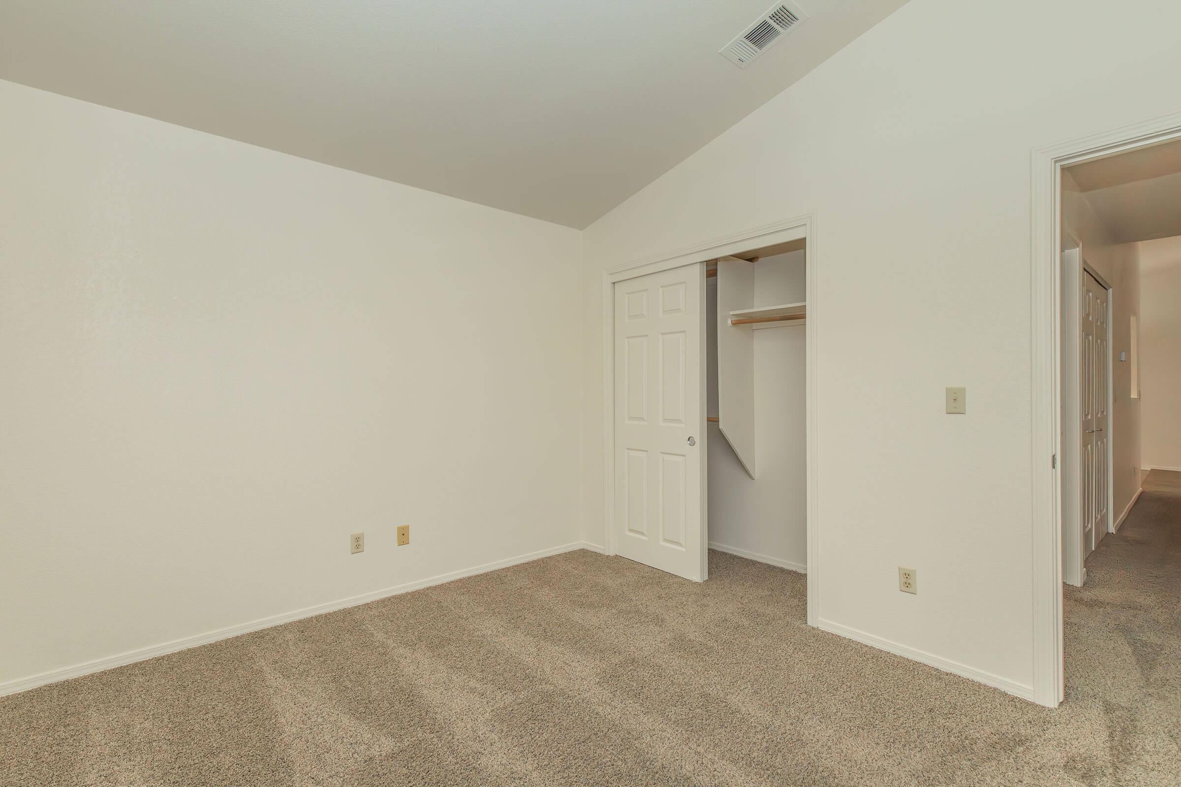 Empty room with light-colored walls and carpet. A closet with white double doors is on the left, while a doorway on the right leads to another room or hallway. The room shows no furniture or decorations, providing a spacious and minimalistic appearance.