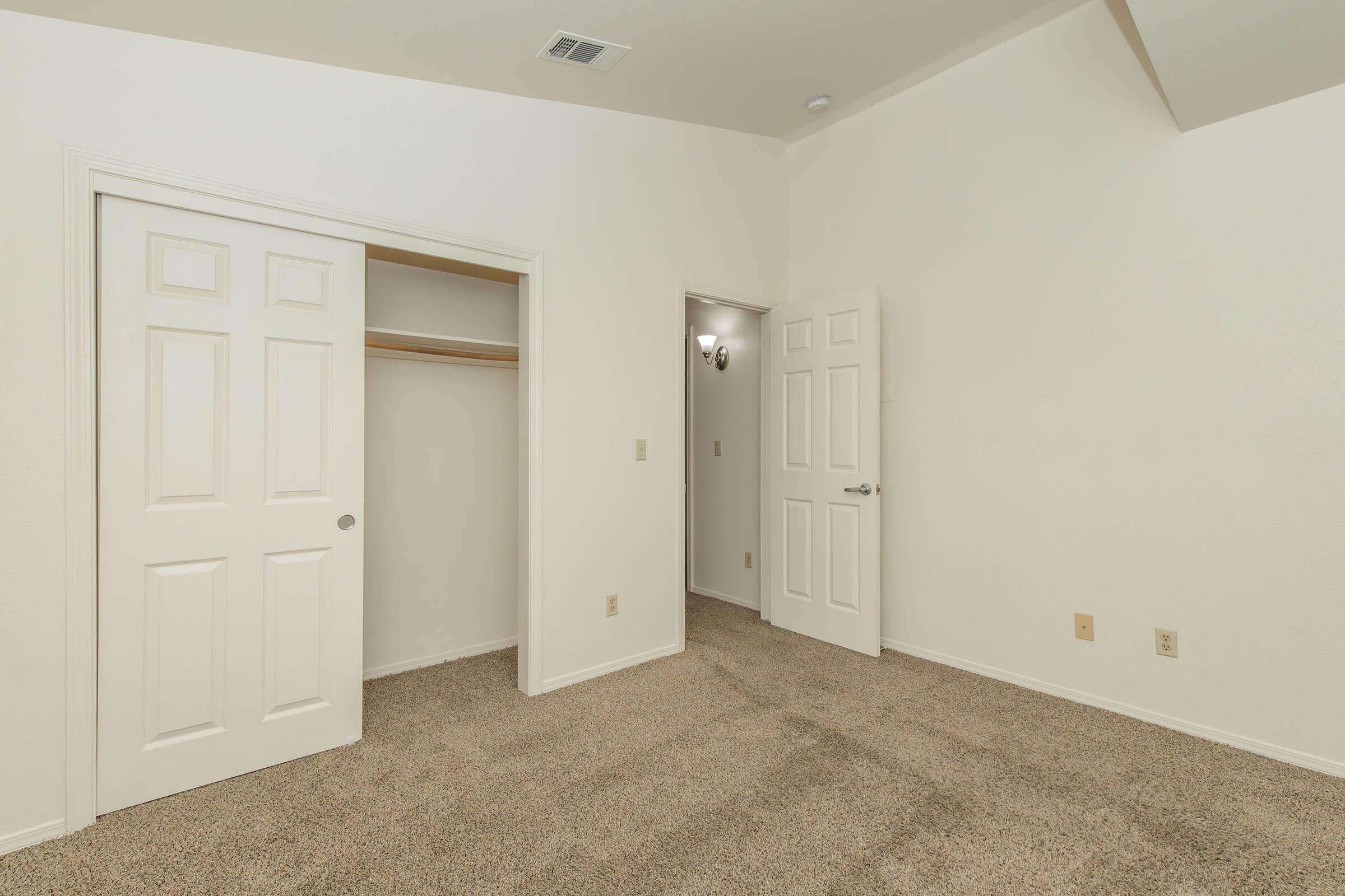 A spacious, empty room with beige carpet. There are two closed white doors: one leading to a closet with wooden shelves and the other to a small hallway. The walls are painted a light color, and the ceiling is plain. Natural light illuminates the space, creating a bright and airy atmosphere.