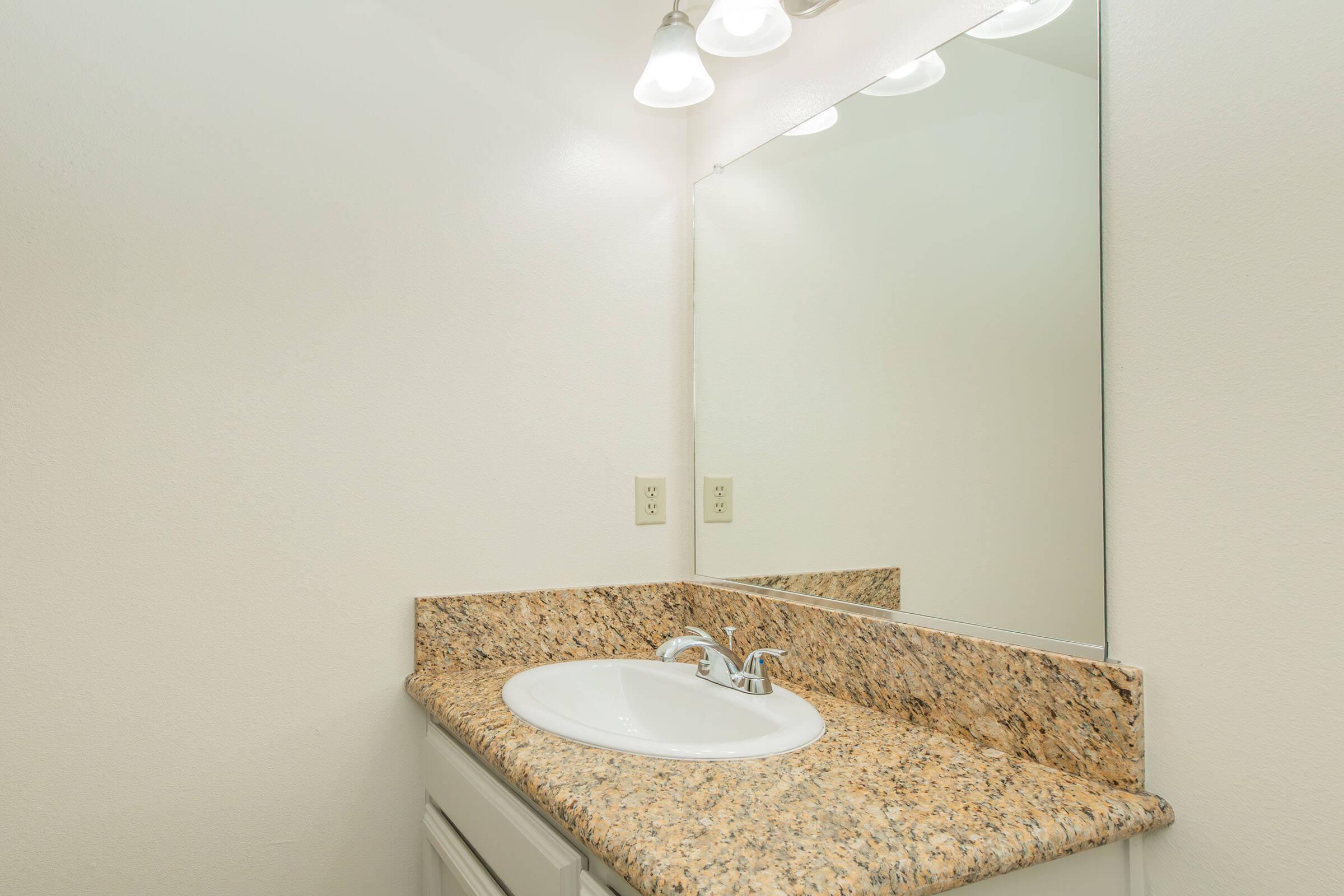 A clean and modern bathroom sink area featuring a granite countertop, a white sink with a chrome faucet, and a large mirror above. Soft lighting from three pendant lights illuminates the space, which has neutral-colored walls, creating a bright and airy atmosphere.