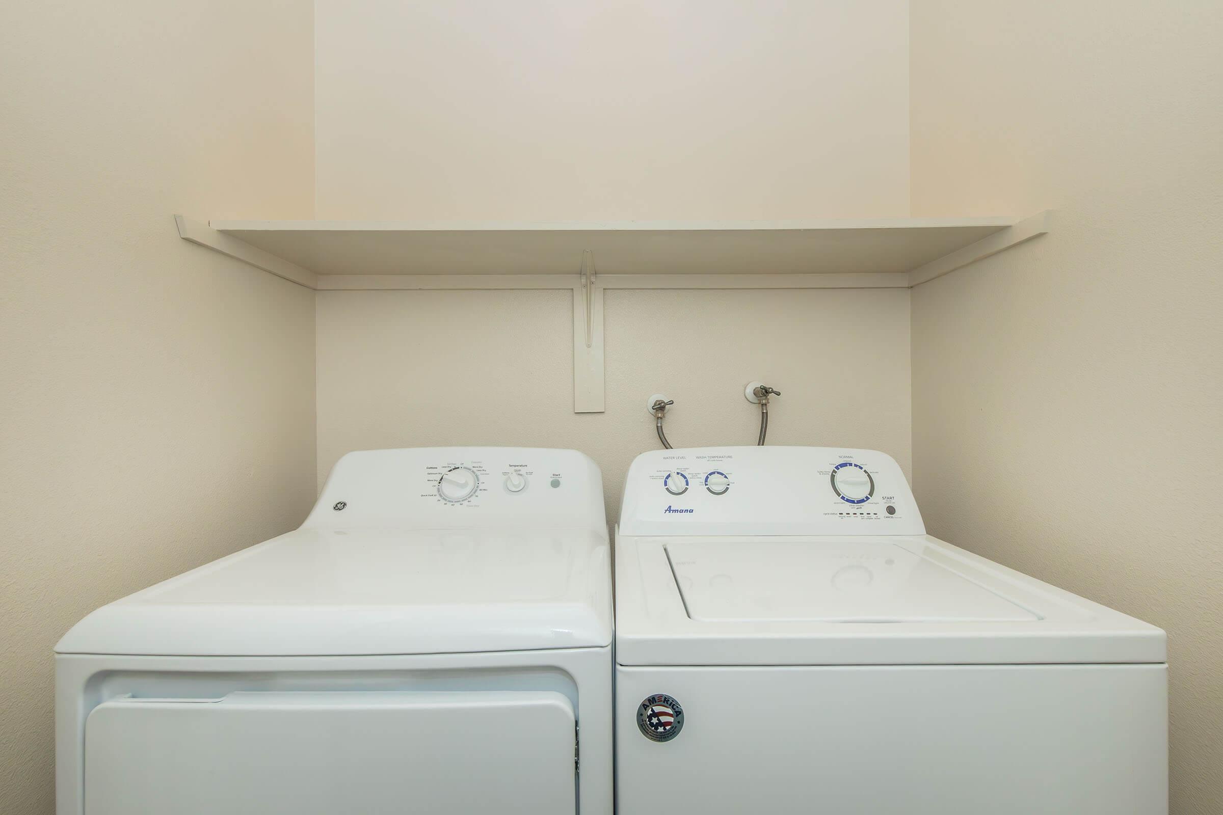 A laundry room featuring a white washing machine and a matching dryer side by side, with a small shelf above them and light-colored walls. The appliances have control dials and water connection points visible.