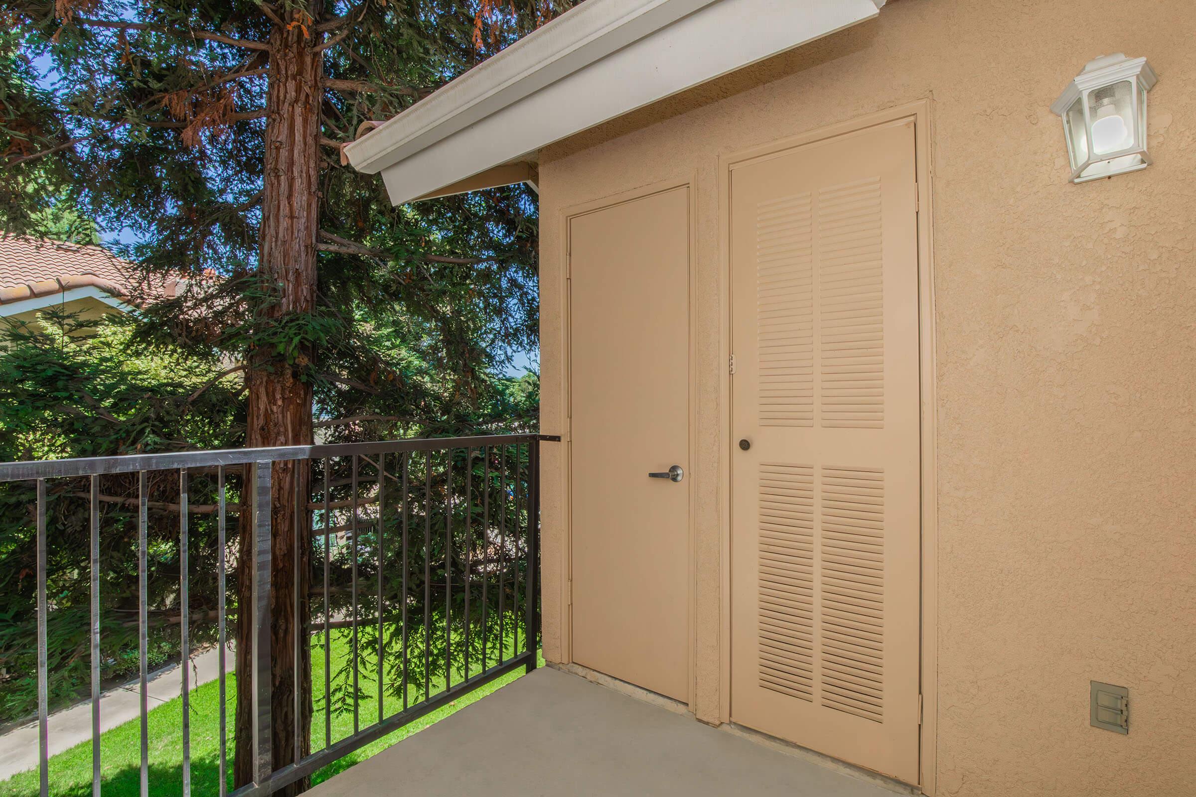 A small balcony area featuring a beige wall, light fixture, and two adjacent doors. Green trees and grass are visible in the background, creating a natural setting. The flooring is plain and the railings are metal.