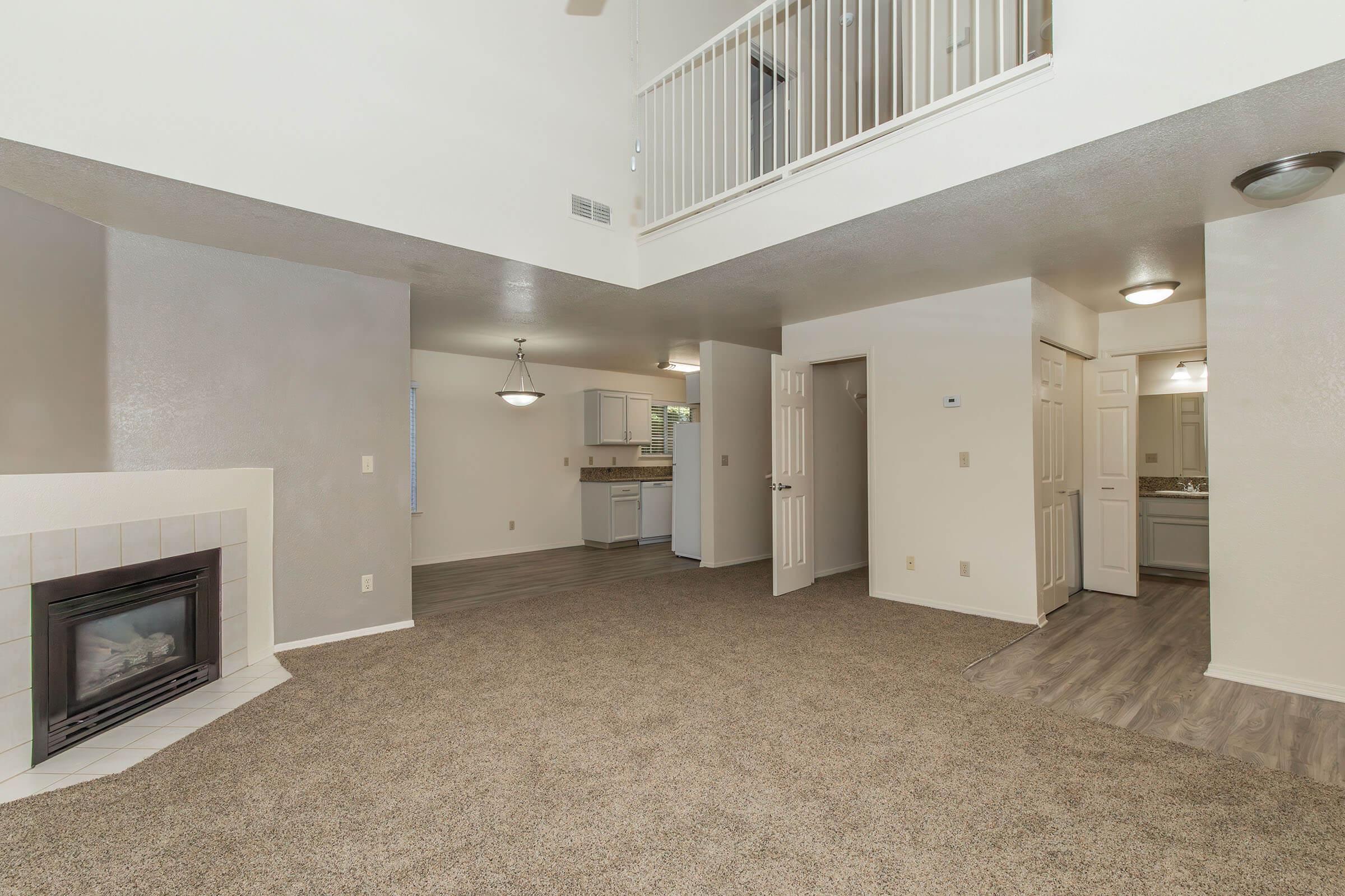Spacious living area with carpet flooring, featuring a corner fireplace. Open layout leading to a bright kitchen area with light-colored cabinets. High ceilings allow for natural light, and a balcony overlooks the space. Doors leading to other rooms are visible, creating an inviting atmosphere.