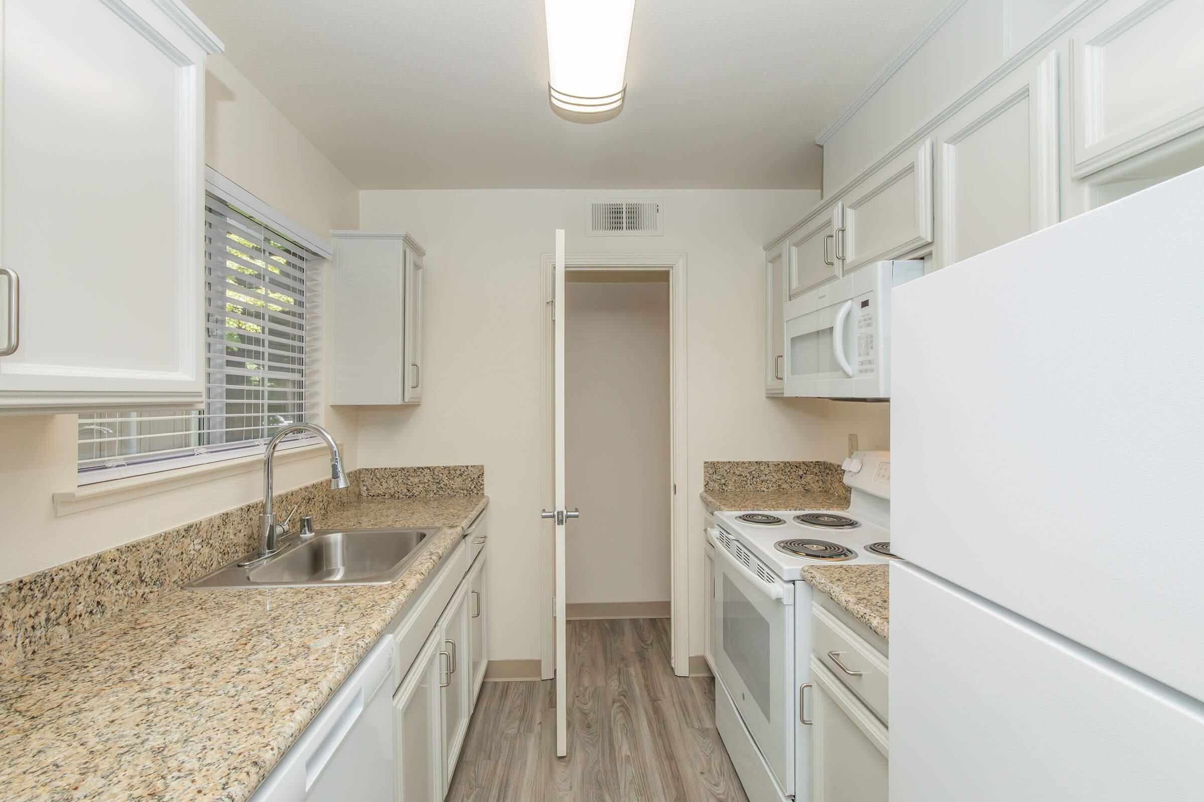 A modern kitchen featuring white cabinetry, a granite countertop, and stainless steel sink. Appliances include a refrigerator, microwave, and stove with an oven. The kitchen has a neutral color scheme with natural light from a window and laminate flooring. A doorway leads to a small adjoining room or pantry.