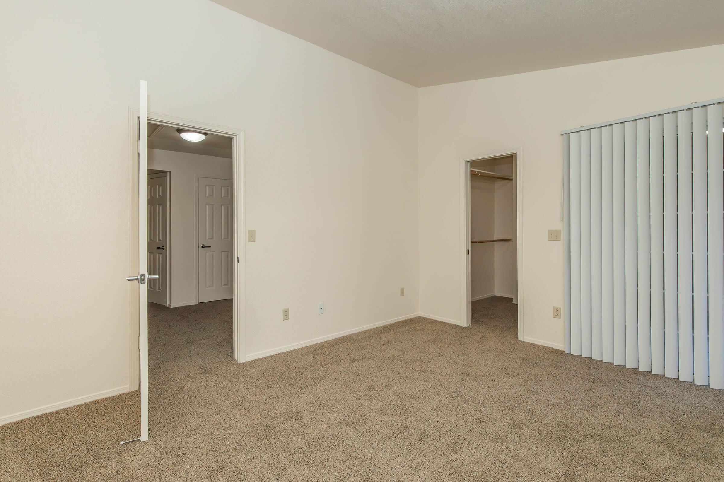 A spacious, empty room with beige carpeting and light-colored walls. A partially open door leads to another room, while a closet is visible in the corner. Vertical blinds cover a window, allowing natural light to enter. The room has a minimalistic design, ideal for various furnishing options.