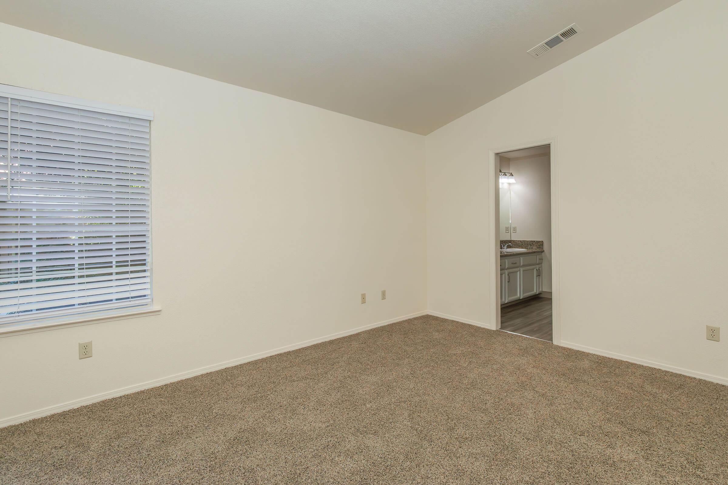A spacious, empty room with beige carpeting and light-colored walls. A window with blinds provides natural light, and a doorway leads to a bathroom area with fixtures visible in the background. The room is uncluttered and ready for furnishing.