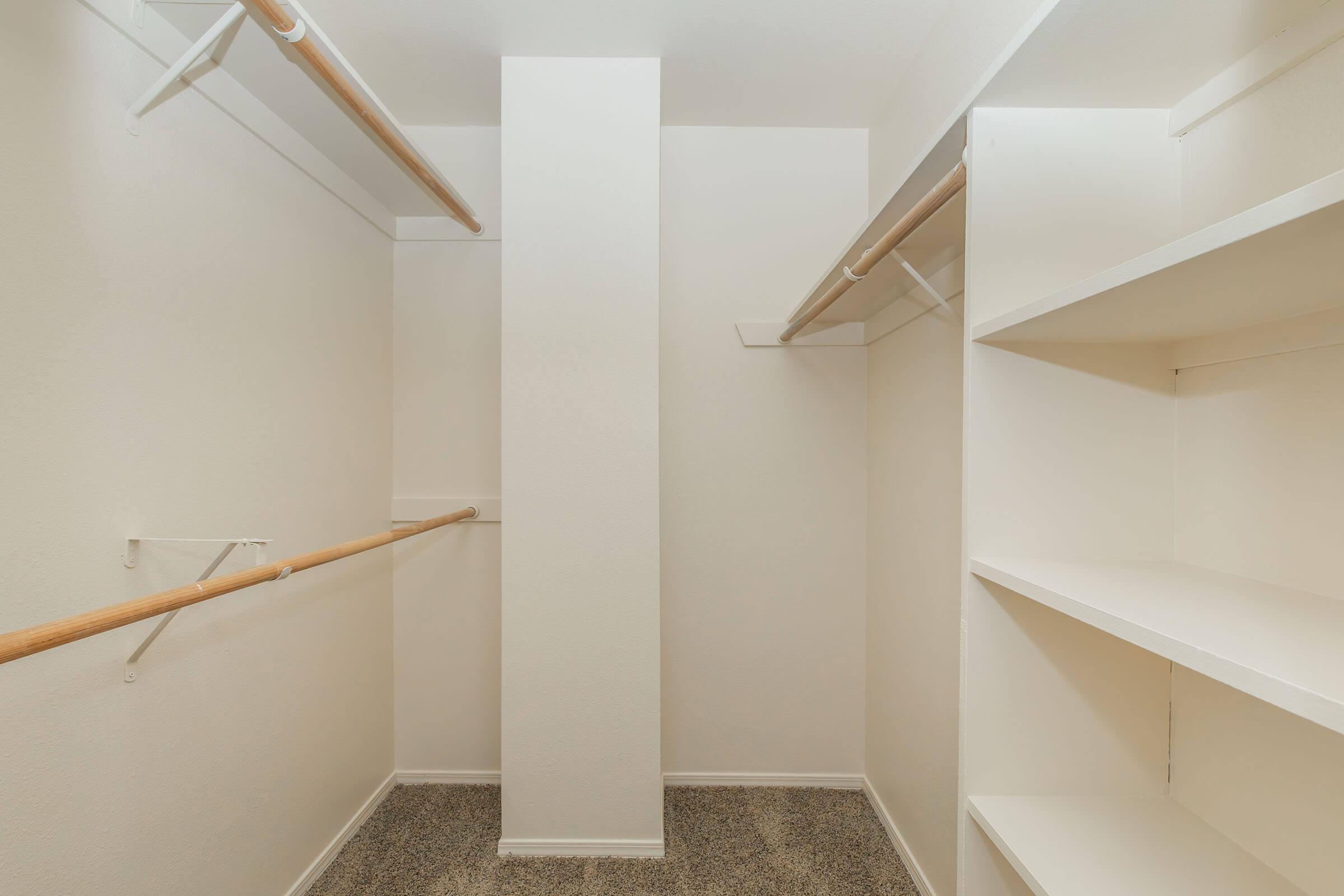 Empty walk-in closet featuring a central support column, wooden hanging rods on either side, and white shelving units. The walls and floors are light-colored, creating a bright and spacious feel. The space is uncluttered and ready for organization or storage.