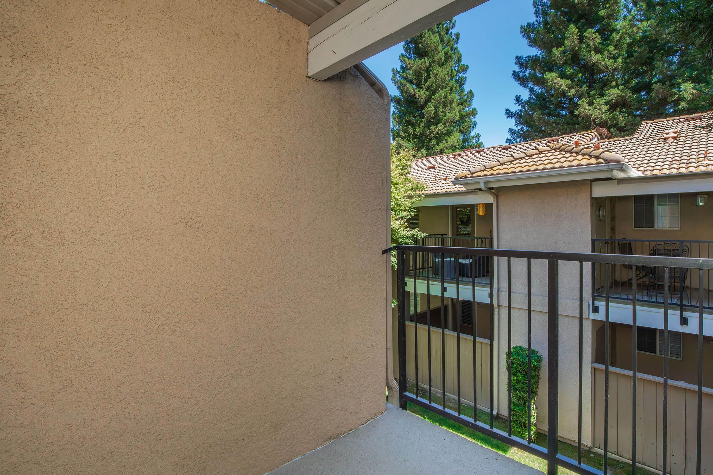 A view from a balcony overlooking a landscaped courtyard with trees and neighboring apartment buildings. The railing is black metal, and the walls are a light beige. Lush greenery adds to the peaceful ambiance of the setting. 