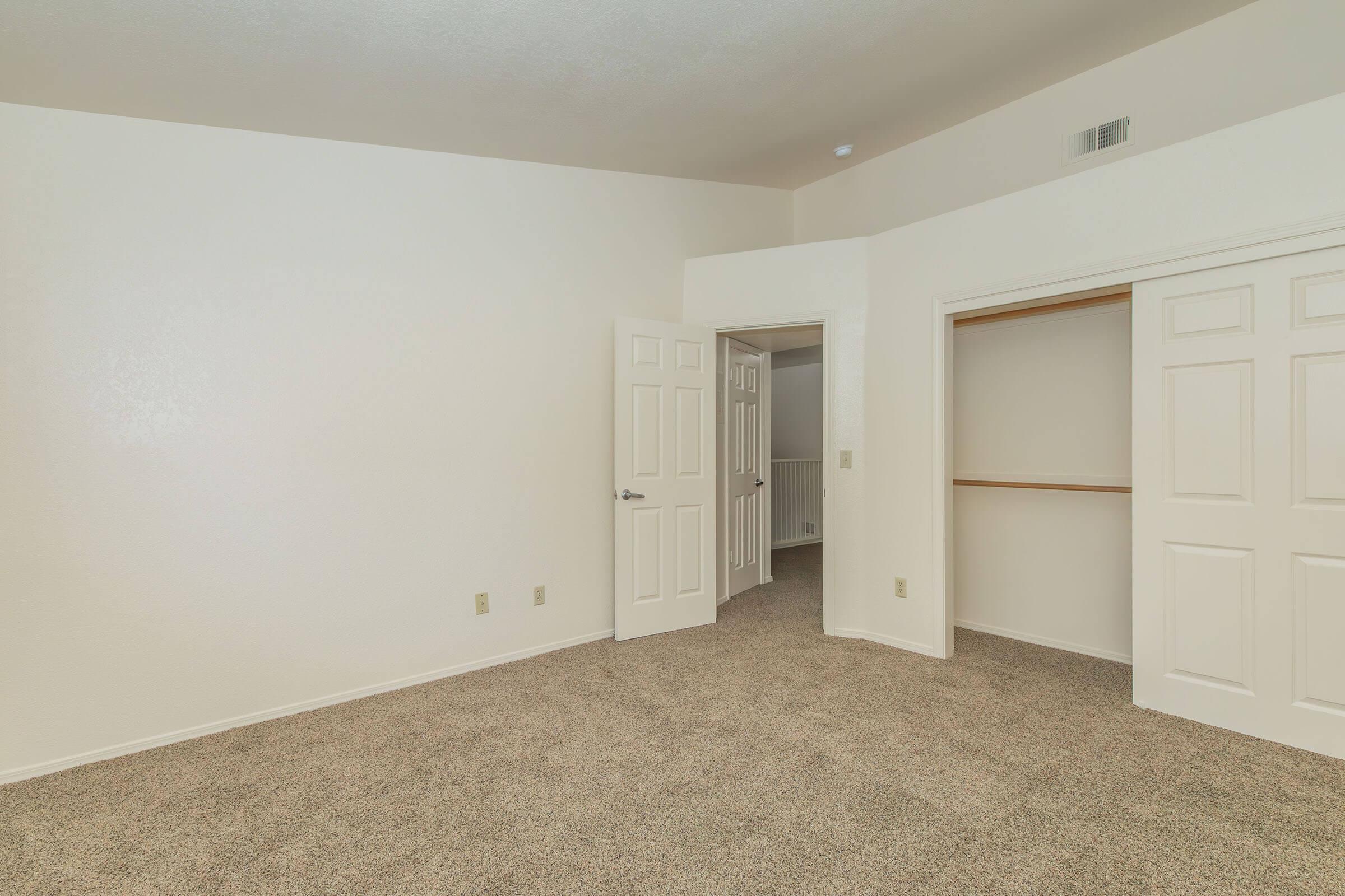 A spacious, empty room with light beige carpet and white walls. Two doors are visible—one leading to a closet and another to an adjacent room. The room has a simple and clean layout, with natural light coming in, creating a bright atmosphere. The ceiling is moderately high, enhancing the sense of openness.