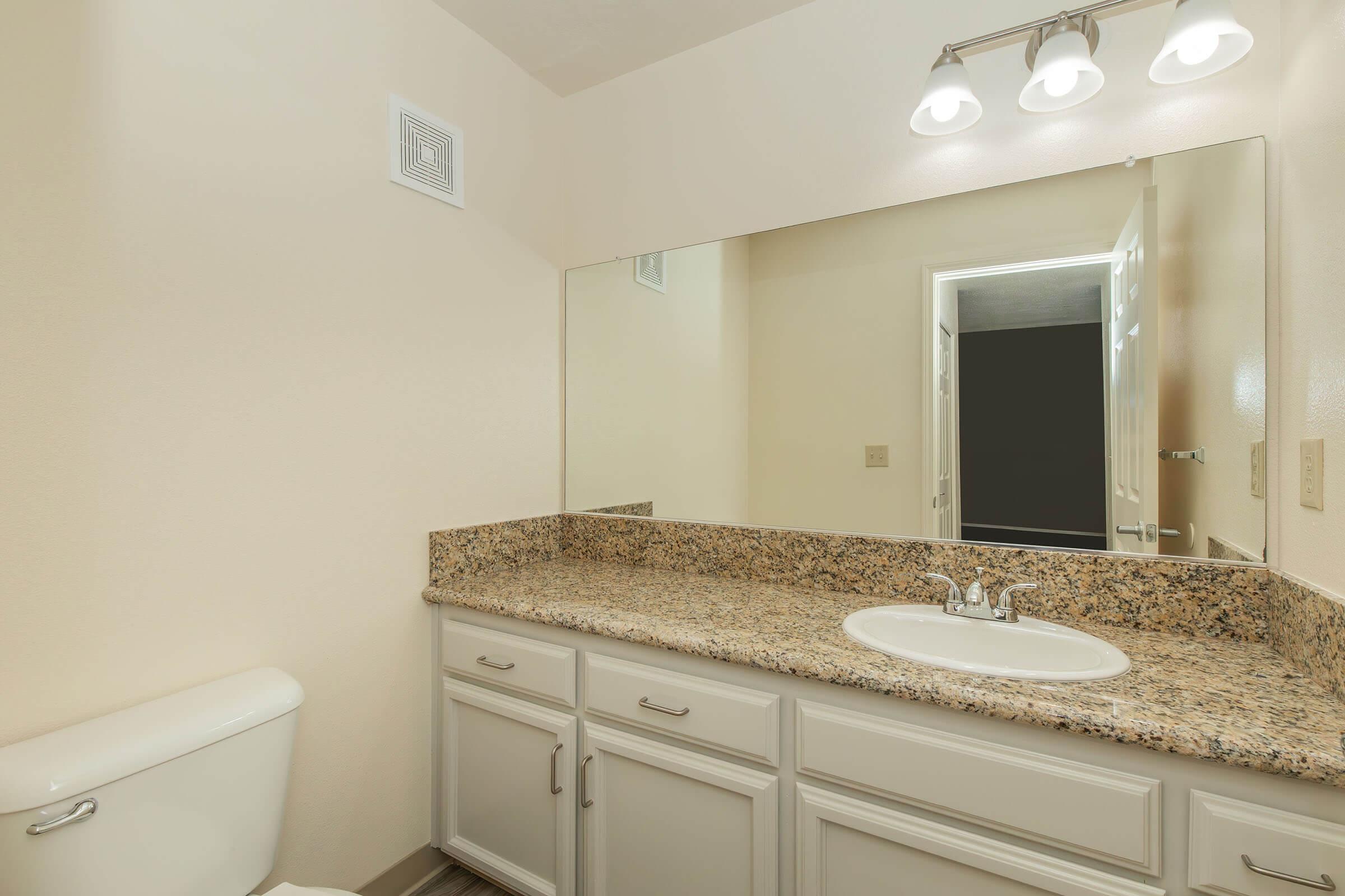A well-lit bathroom featuring a granite countertop with a sink, white cabinetry, a toilet, and a large mirror. The walls are a light shade, creating a spacious feel, and there is a door leading to another room in the background.