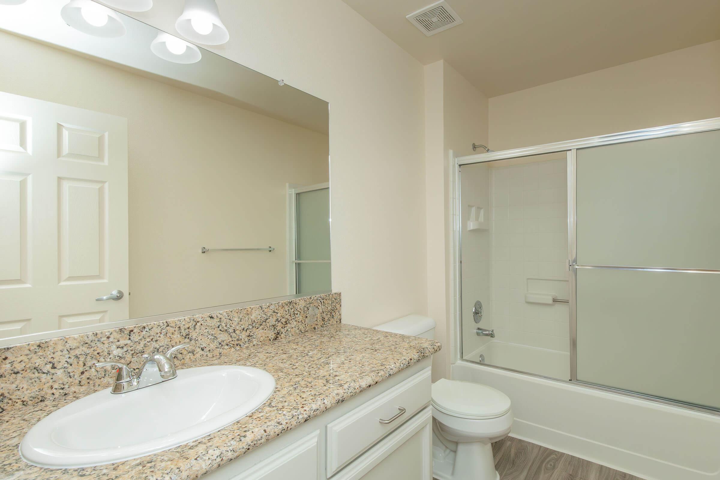 A modern bathroom featuring a light-colored granite countertop with a sink, a large mirror, a glass shower enclosure, a white toilet, and neutral-toned walls and flooring. Natural light brightens the space, creating a clean and spacious atmosphere.