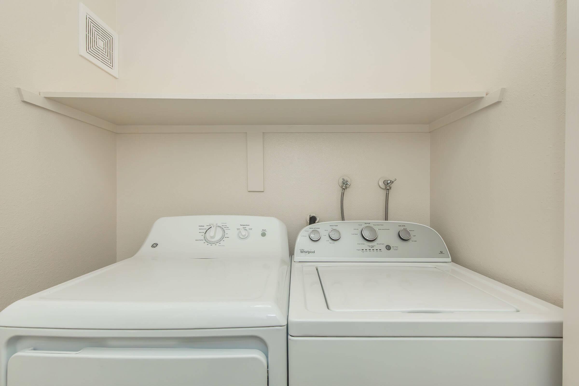 A washer and dryer side by side in a laundry room. The washer is on the left and the dryer is on the right, both are white and modern. Above them, there's a shelf mounted on the wall. The walls are painted a light color, providing a clean and organized appearance.
