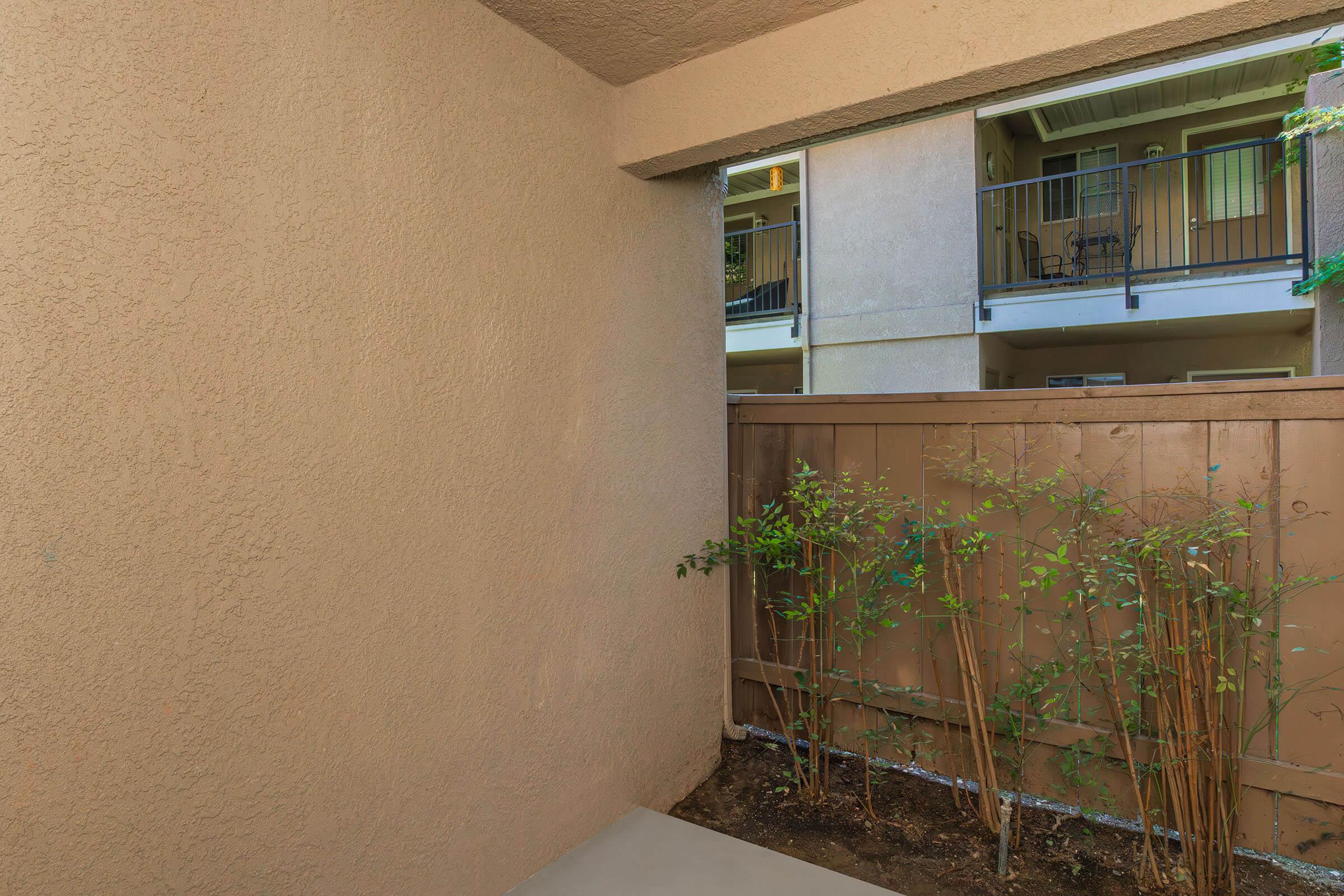 View of a small outdoor space with a beige wall and a fence. In the background, there are balconies of nearby apartments, with a few green plants growing near the fence. The scene is well-lit, suggesting a bright atmosphere.