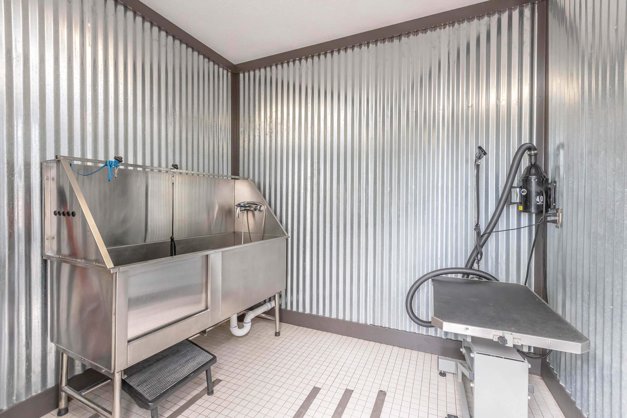 A clean, industrial-style grooming room featuring a stainless steel bathtub with a faucet, a grooming table with an adjustable arm, and silver corrugated metal walls. The space is well-lit and tiled, designed for pet grooming services.