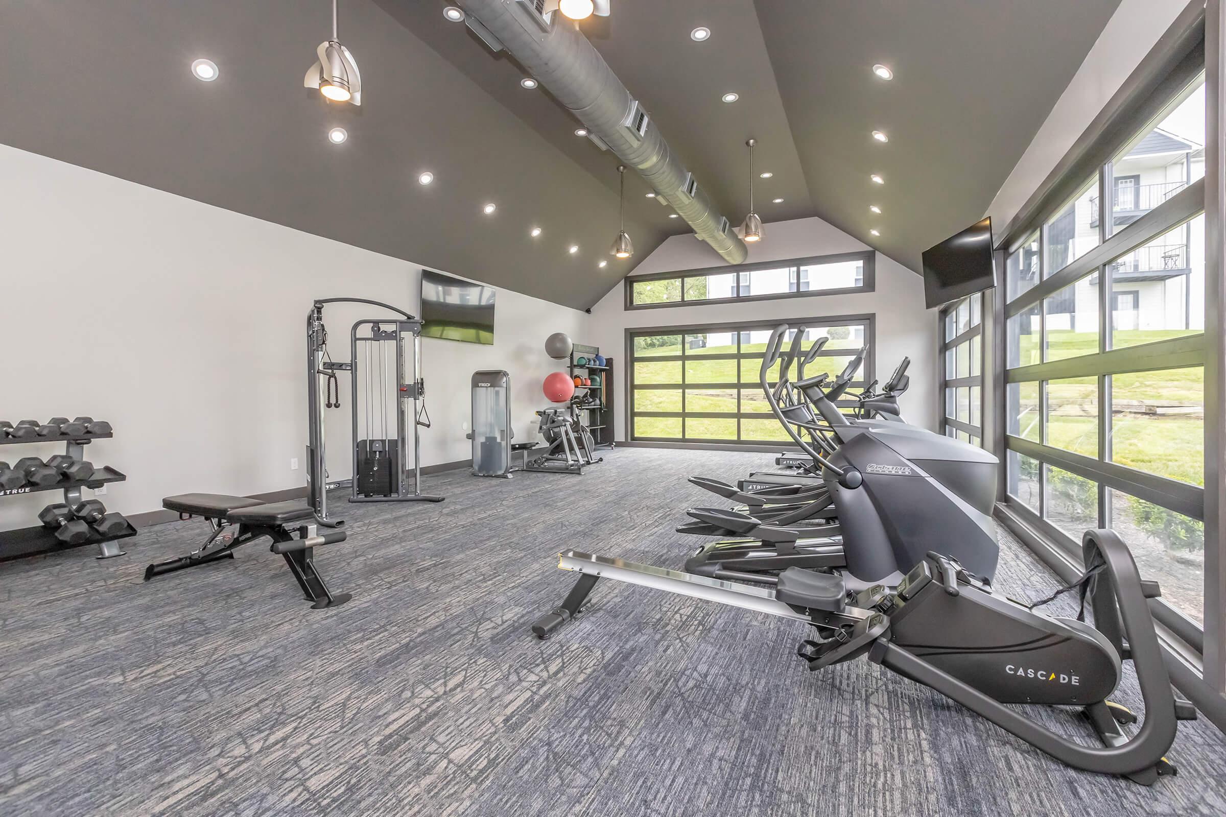 A modern gym interior featuring various exercise equipment, including elliptical machines, a weight bench, and free weights. The space has large windows for natural light, a dark ceiling with recessed lighting, and a clean, contemporary aesthetic.