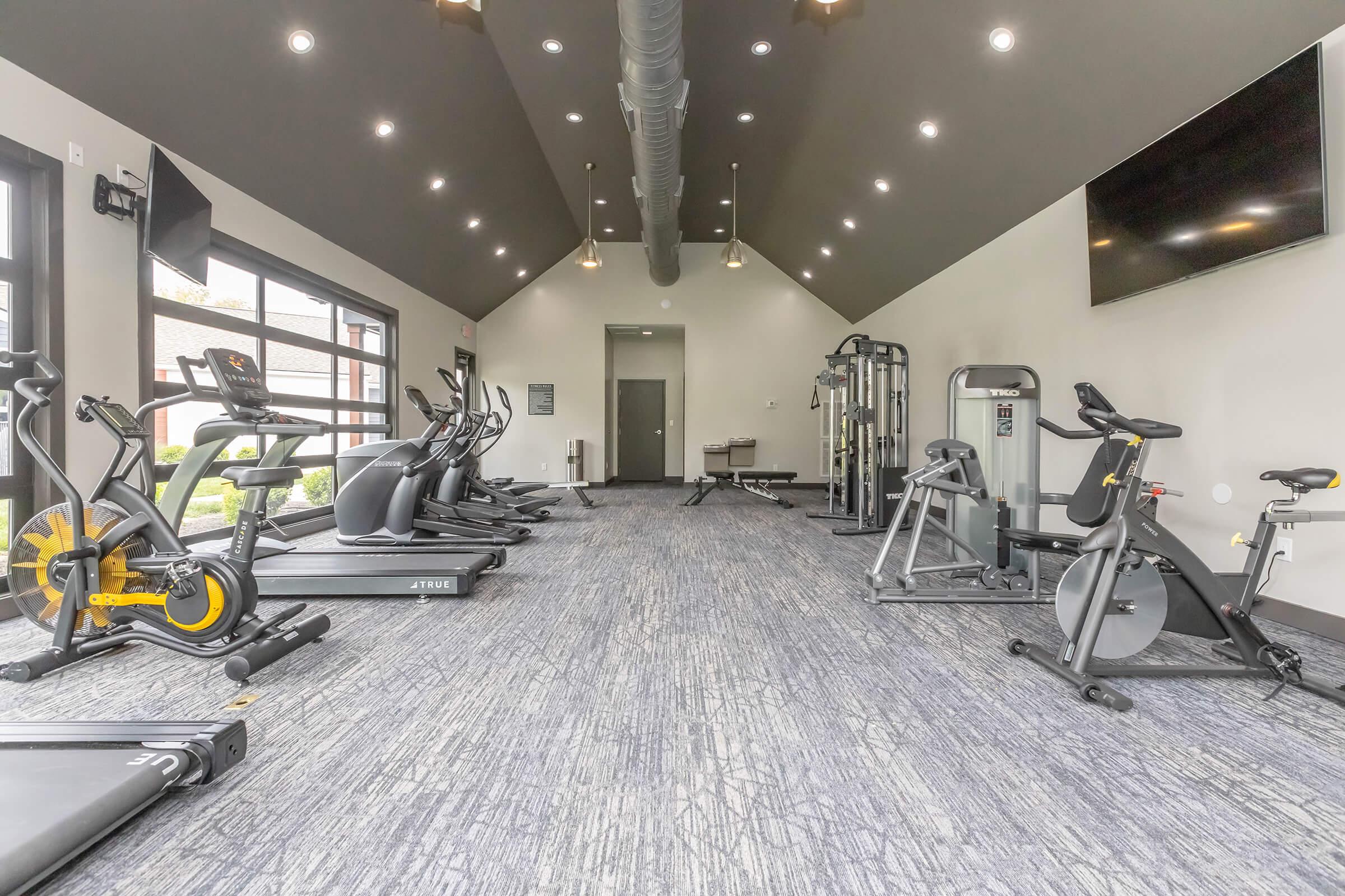 A modern fitness room featuring various exercise equipment, including treadmills, stationary bikes, ellipticals, and weight machines. The space has a high ceiling with recessed lighting, large windows allowing natural light, and a flat-screen TV mounted on the wall.