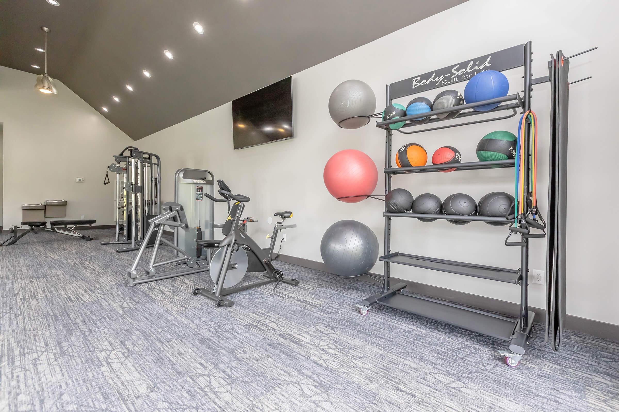 A modern gym interior featuring various fitness equipment, including a stationary bike, resistance machines, and a rack of stability balls and resistance bands. The room has a light color scheme and is well-lit with overhead lighting and a wall-mounted TV.