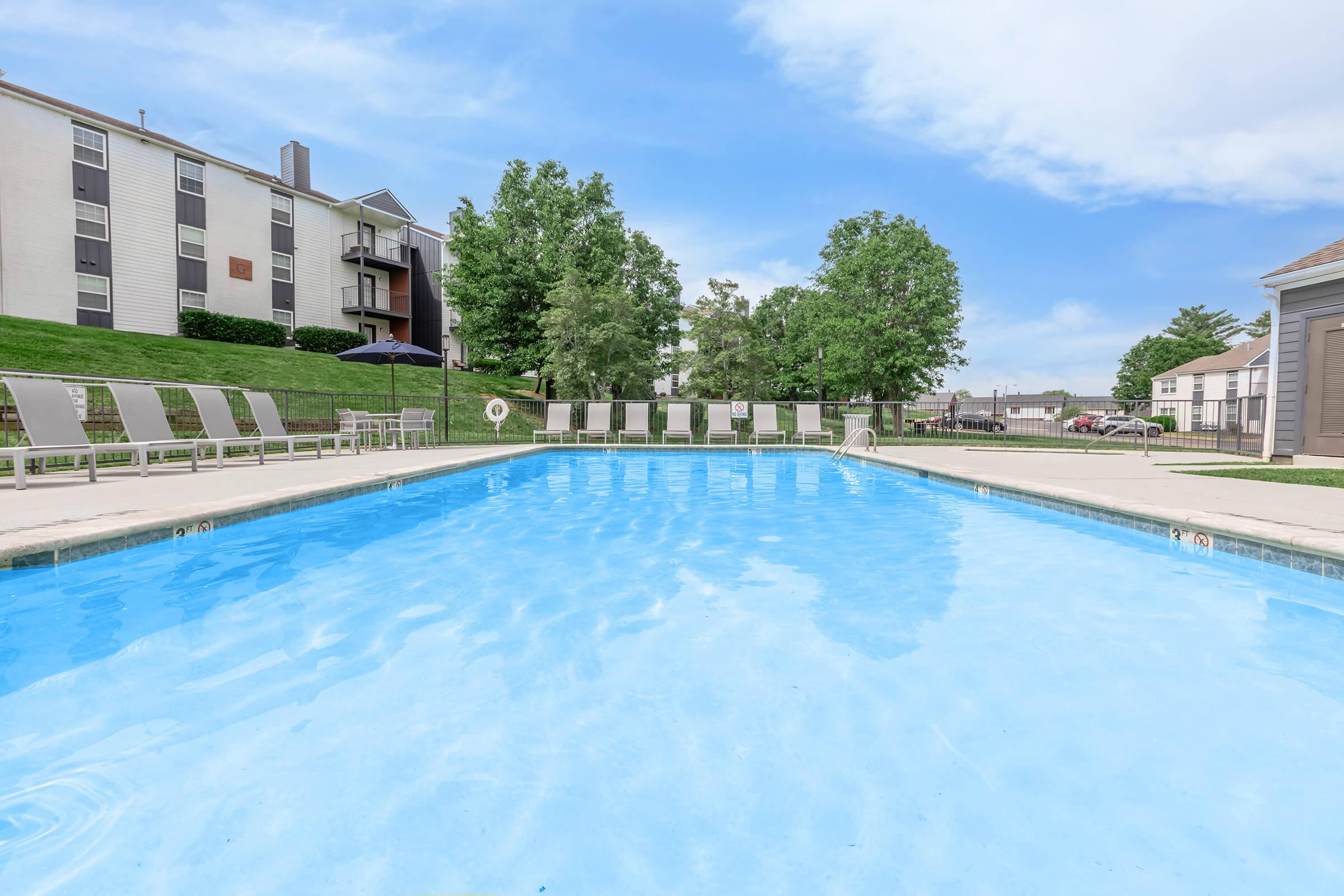 A clear swimming pool surrounded by lounge chairs and green trees. The pool features a well-maintained deck and a nearby building, creating a relaxing outdoor space for leisure and recreation. The sky is partly cloudy, adding to the serene atmosphere.