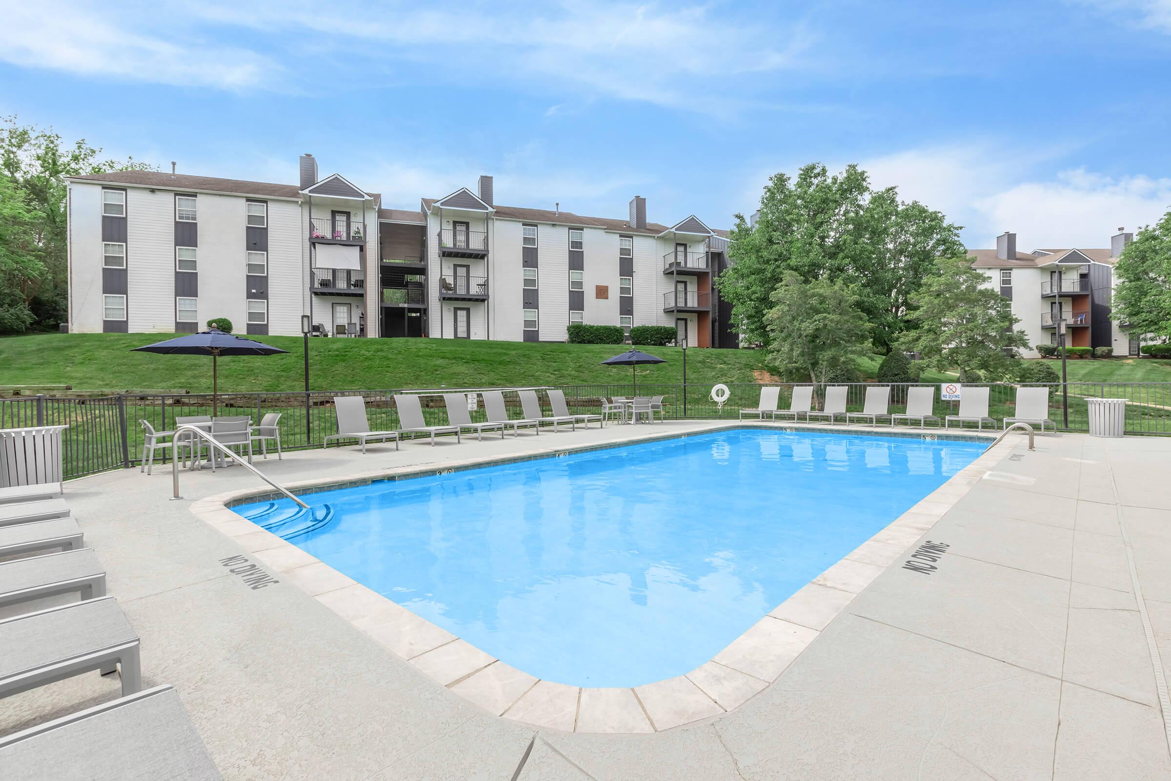A clear swimming pool surrounded by tan lounge chairs, with umbrellas, against a backdrop of green grass and multi-story apartment buildings. The sky is bright with a few clouds, creating a tranquil and inviting atmosphere for relaxation and leisure.
