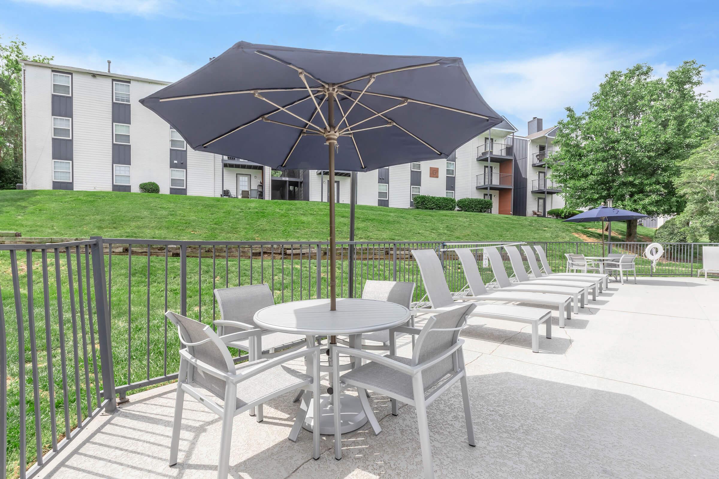A patio area featuring a round table with chairs under a large umbrella, surrounded by lounge chairs. In the background, there are grassy areas and residential buildings with balconies, set against a clear blue sky.