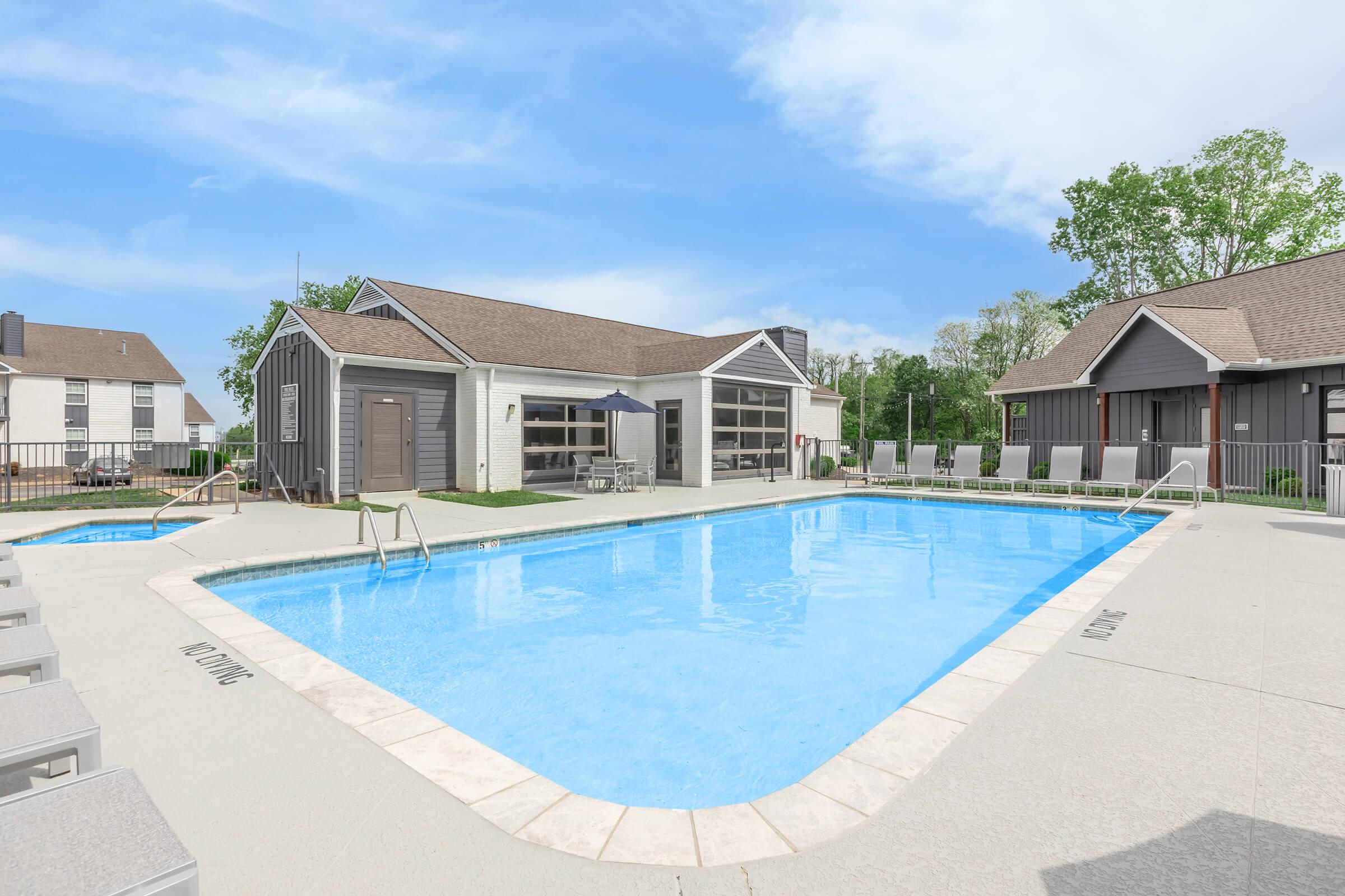 A clear blue swimming pool surrounded by a deck with lounge chairs. In the background, there are buildings, including a clubhouse with large windows and outdoor seating. Lush greenery and a bright blue sky enhance the inviting atmosphere of this outdoor space.