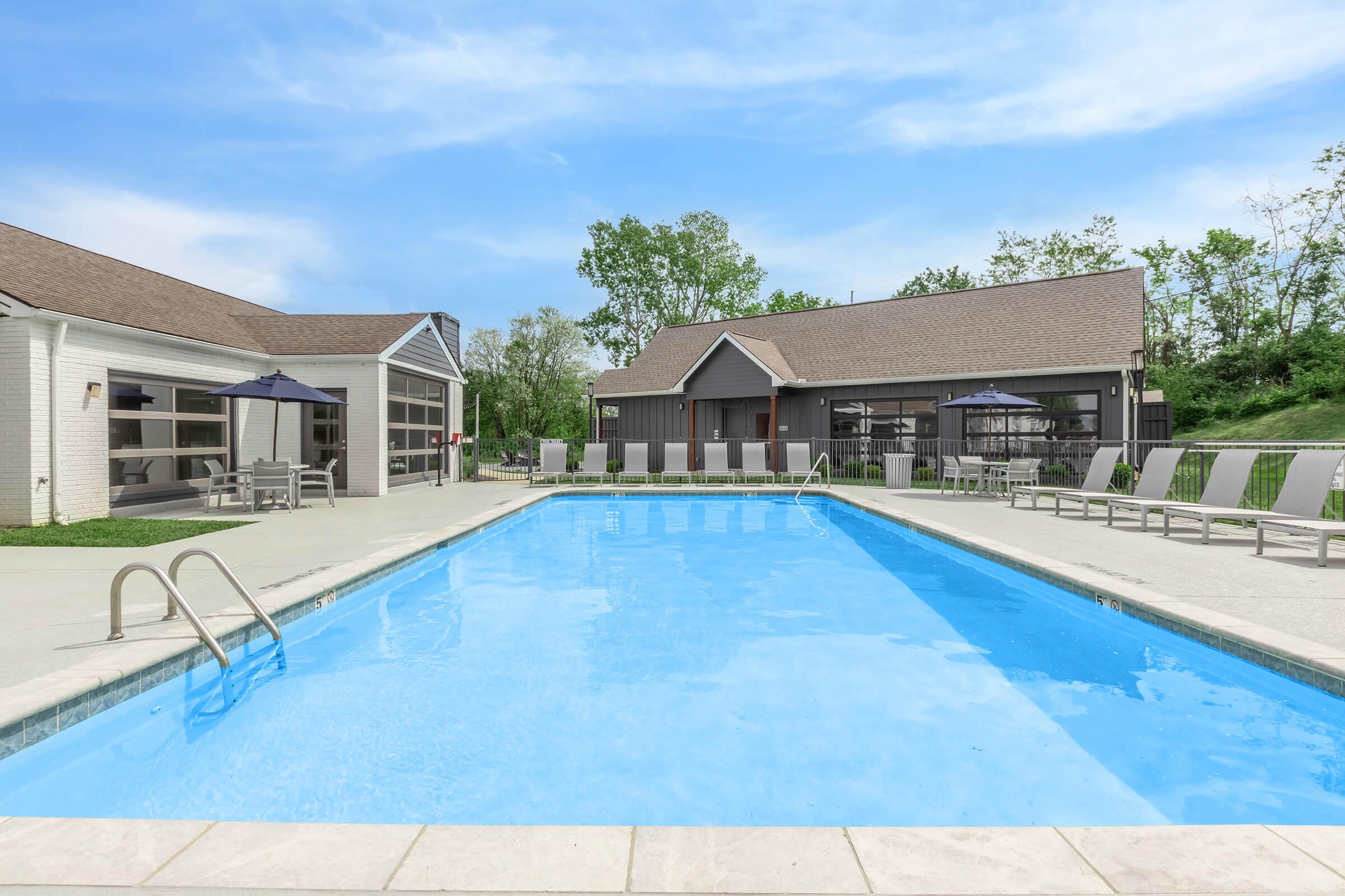 A clear blue swimming pool surrounded by lounge chairs and umbrellas, with green trees in the background. The area features modern buildings alongside the pool, creating a relaxing outdoor space for leisure and relaxation.