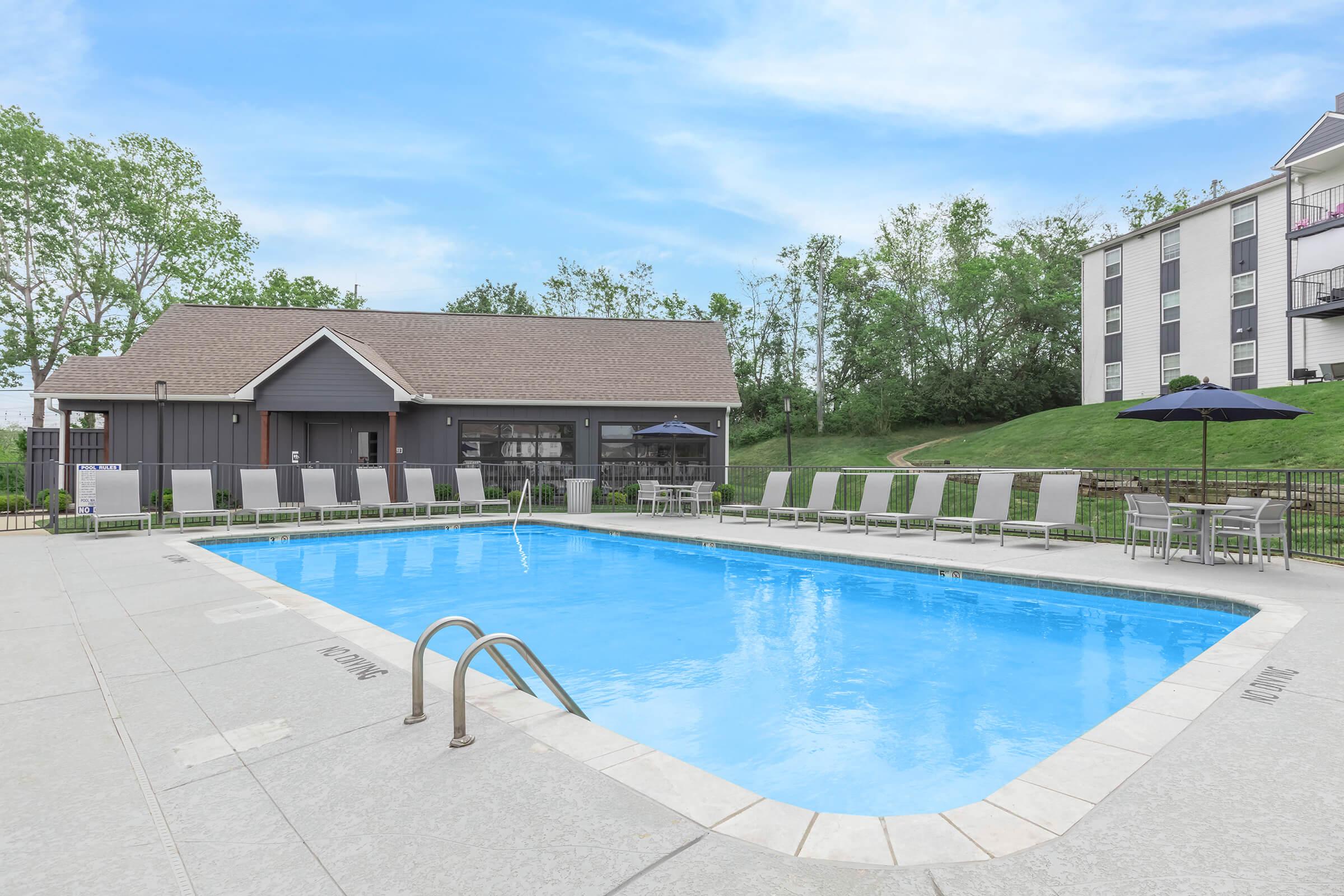 A clear blue swimming pool surrounded by lounge chairs and tables. There are shaded umbrellas near the pool, and a building with a sloped roof is visible in the background. Lush greenery surrounds the area, under a bright blue sky.