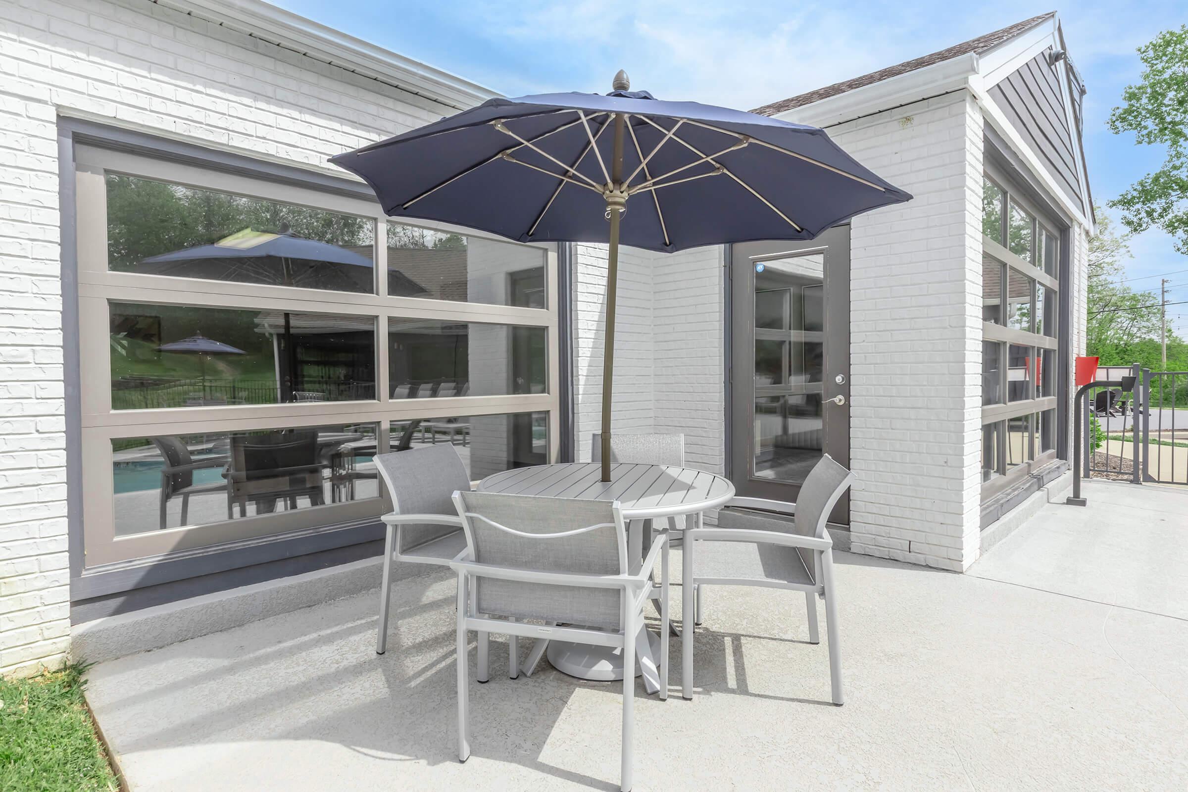 A modern outdoor seating area featuring a round table and six chairs under a navy blue umbrella. The setting is adjacent to a white-brick building with large windows, reflecting a clear blue sky. Green grass borders the patio, creating a relaxing and inviting outdoor space.