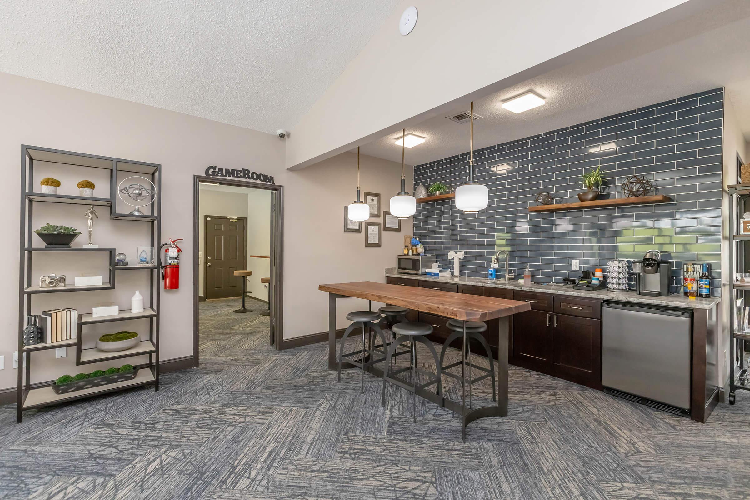 Modern community kitchen area featuring a sleek design with a wooden dining table and bar stools, glass storage shelves displaying decorative items, and a dark tiled backsplash. The space includes appliances like a refrigerator and coffee maker, with soft lighting fixtures enhancing the inviting atmosphere.