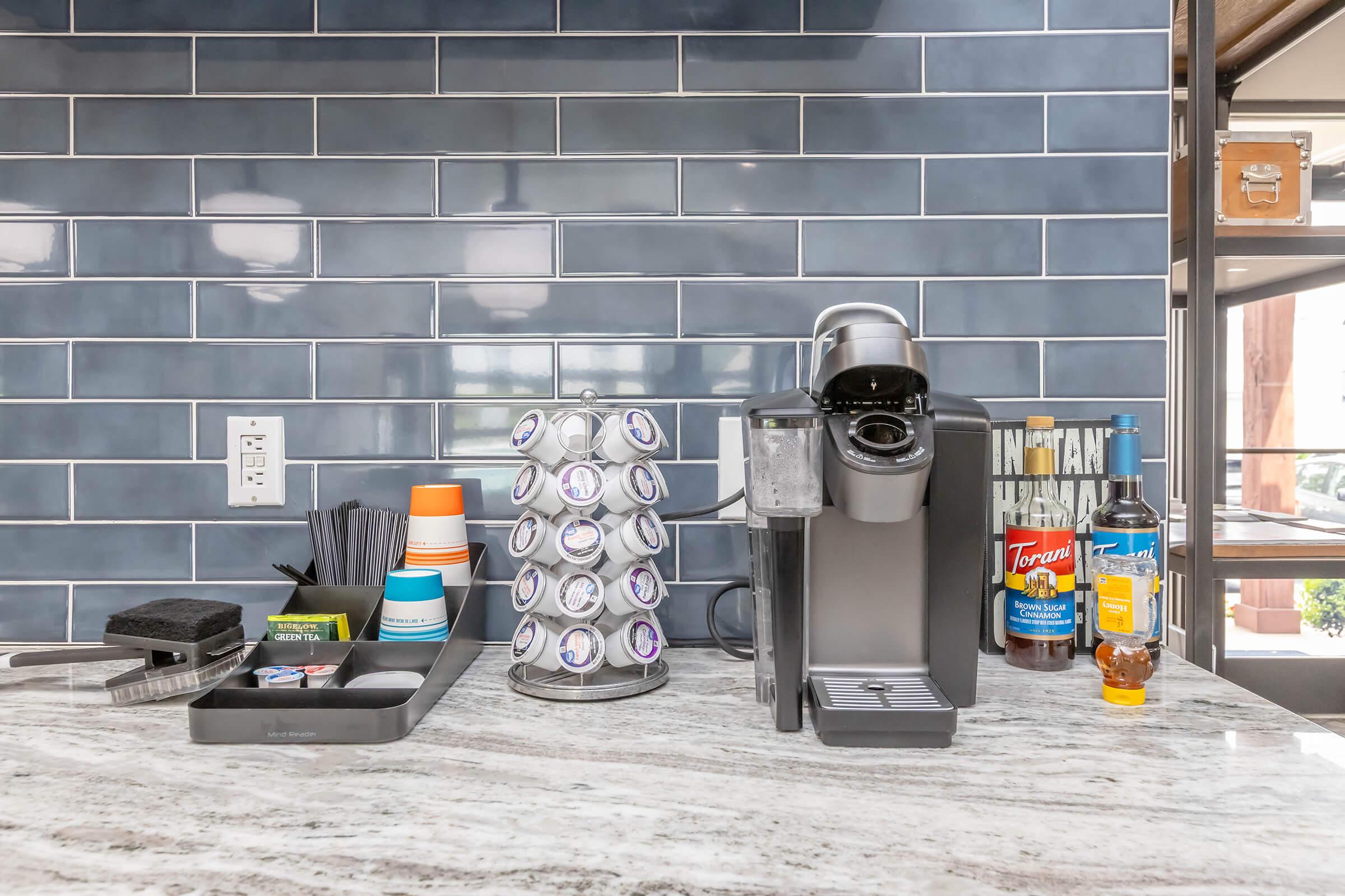A kitchen countertop featuring a coffee machine with a pod holder stacked with coffee pods. Nearby are various condiments including sugar, creamers, and a bottle of syrup, alongside utensils in a tray and a sponge. The backsplash has blue tiles, and the overall setting is bright and organized.
