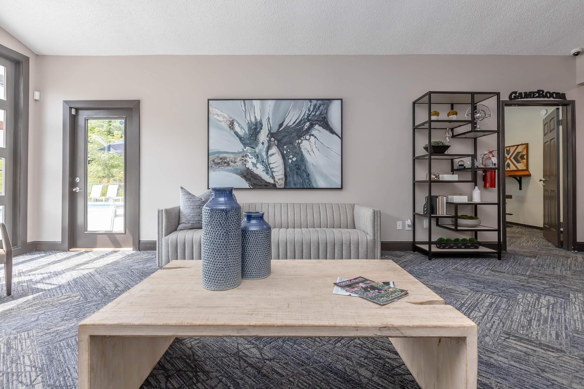 A modern interior space featuring a light gray sofa, a wooden coffee table, and decorative vases. There's an abstract painting on the wall and a bookshelf showcasing decorative items. A door leads to another room, and large windows let in natural light, creating an inviting atmosphere.
