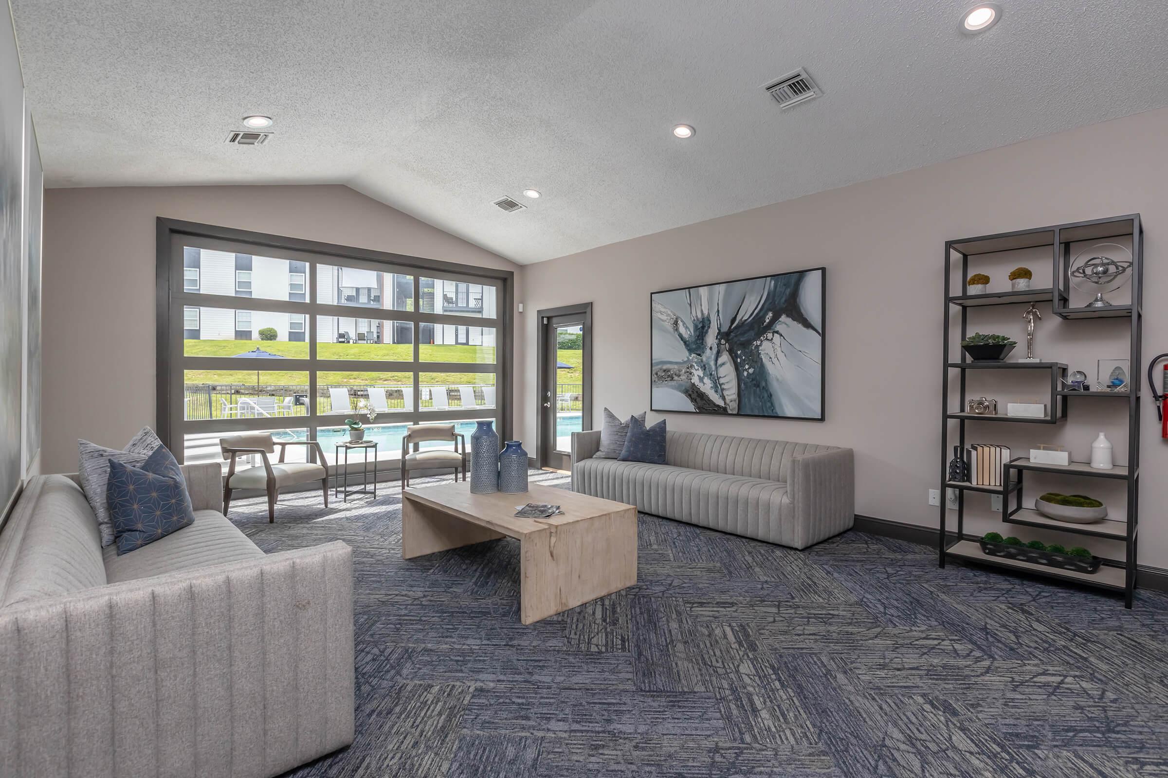 A modern lounge area with two light gray sofas, a wooden coffee table, and decorative elements. Large windows provide natural light and a view of an outdoor pool area. The walls are painted in neutral tones, and there's a stylish shelf displaying various decor items.