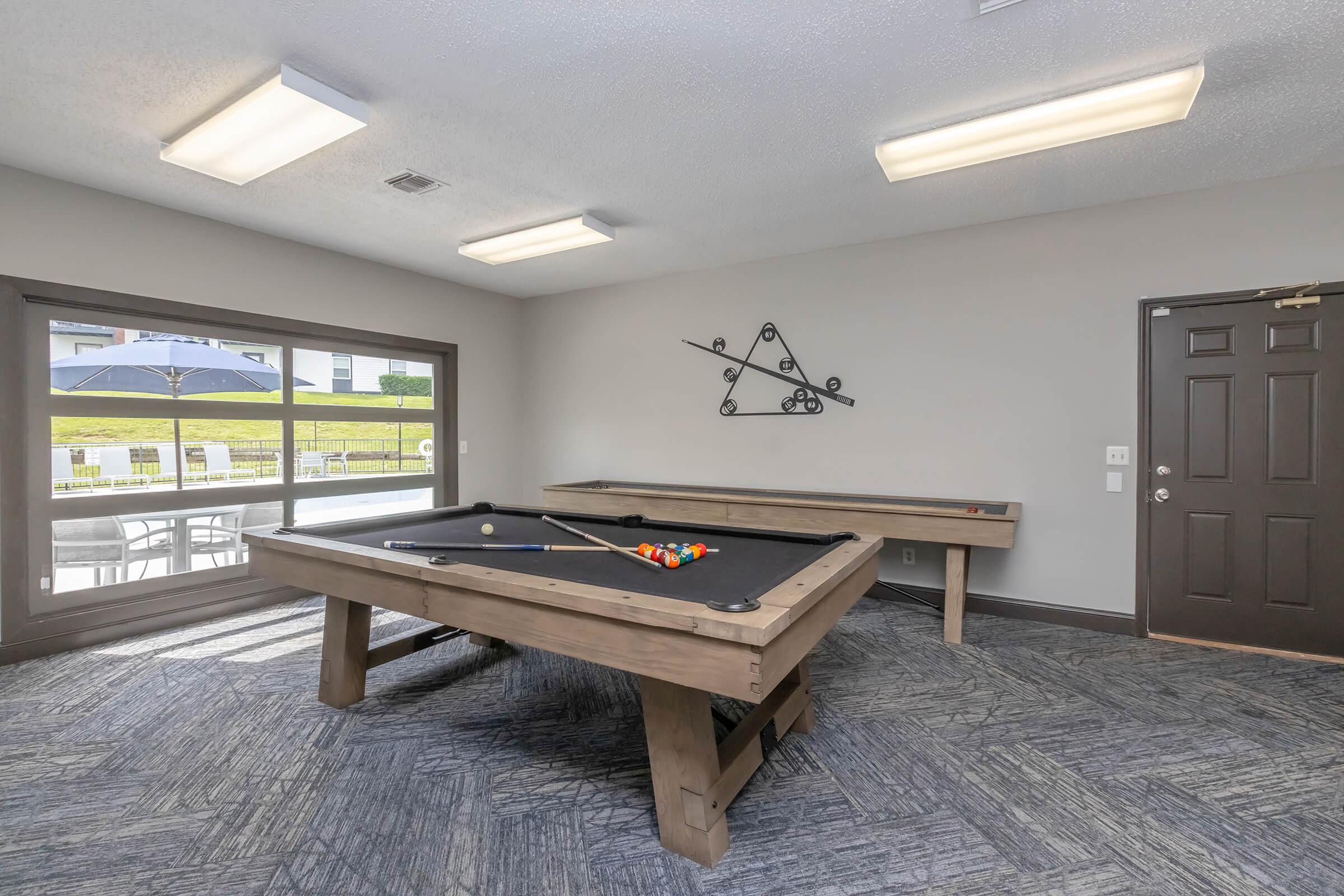 An indoor recreational area featuring a pool table with colorful billiard balls, a seating area near large windows, and modern lighting. The walls are adorned with minimalist decor, creating a relaxing atmosphere for leisure activities.