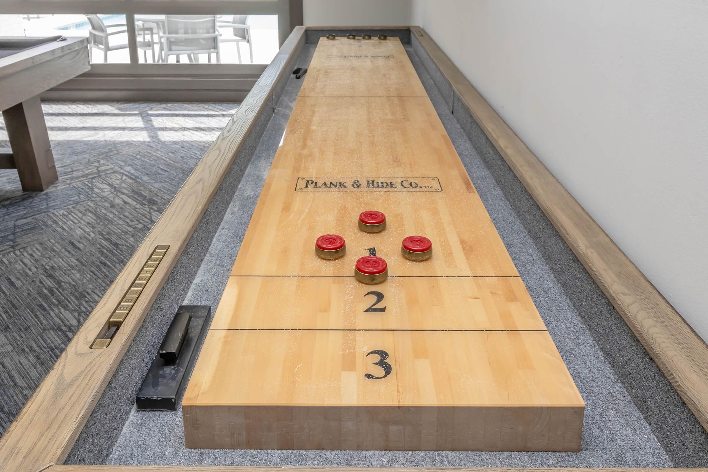 A shuffleboard table with a wooden playing surface, featuring three red discs positioned on the board. The table has markings indicating scoring zones and the brand "Plank & Hide Co." is engraved on it. The background shows a spacious room with additional furniture.