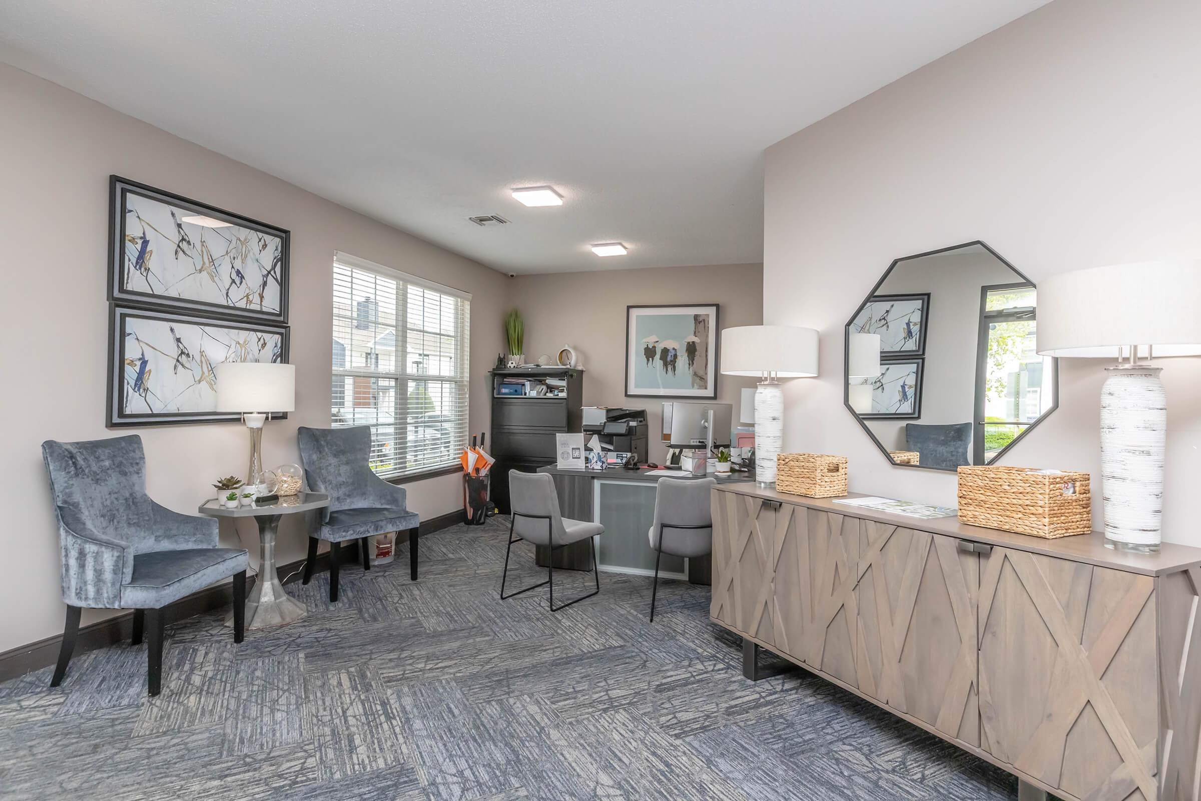 A modern office space featuring two gray chairs and a round table. There’s a desk with a computer, decorative items, and a mirror on the wall. The room is well-lit with large windows and has artwork displayed, creating an inviting and professional atmosphere.