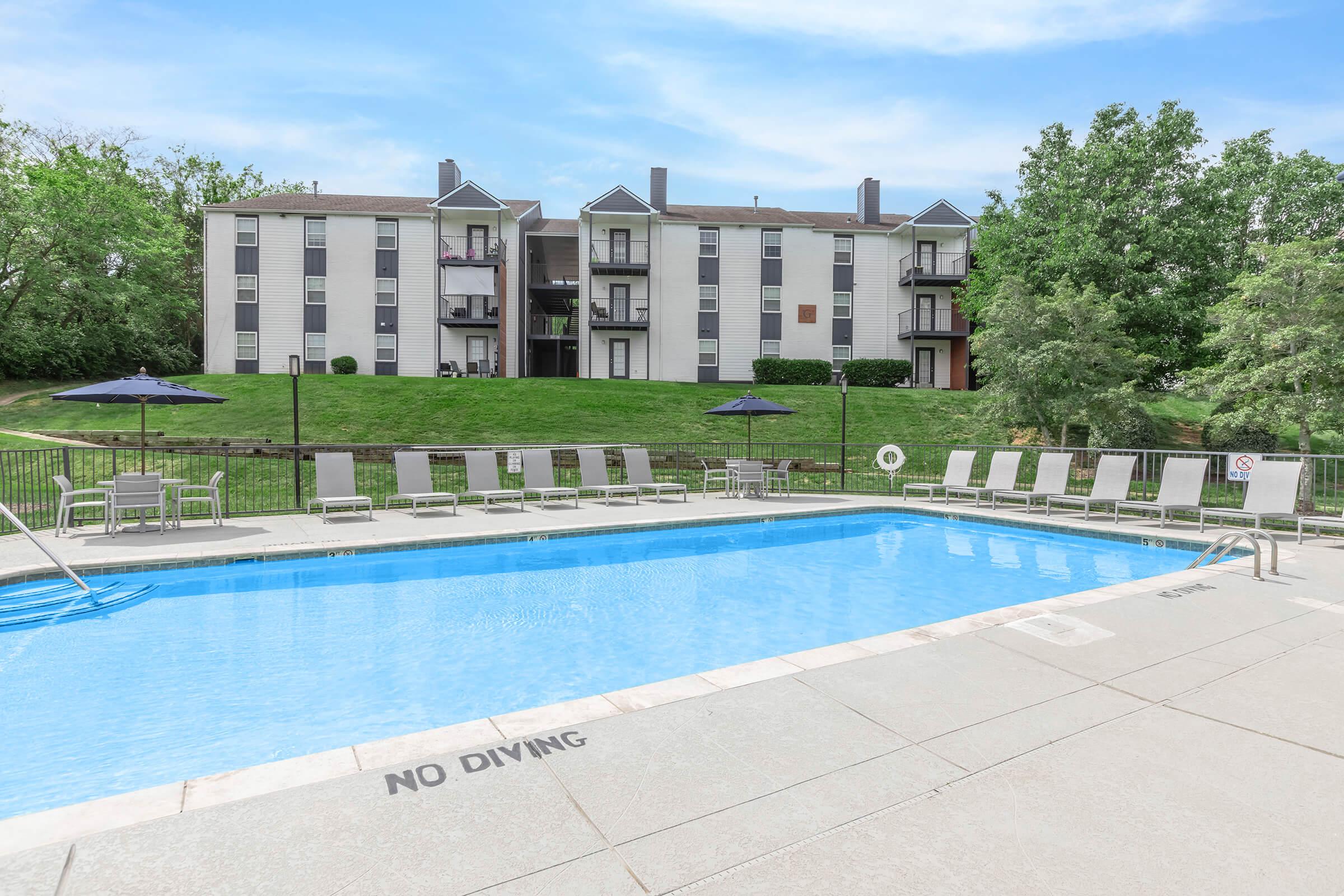 A clear swimming pool surrounded by lounge chairs and umbrellas, with a grassy area and apartments in the background. The pool deck has a "No Diving" sign, and the sky is bright with few clouds.
