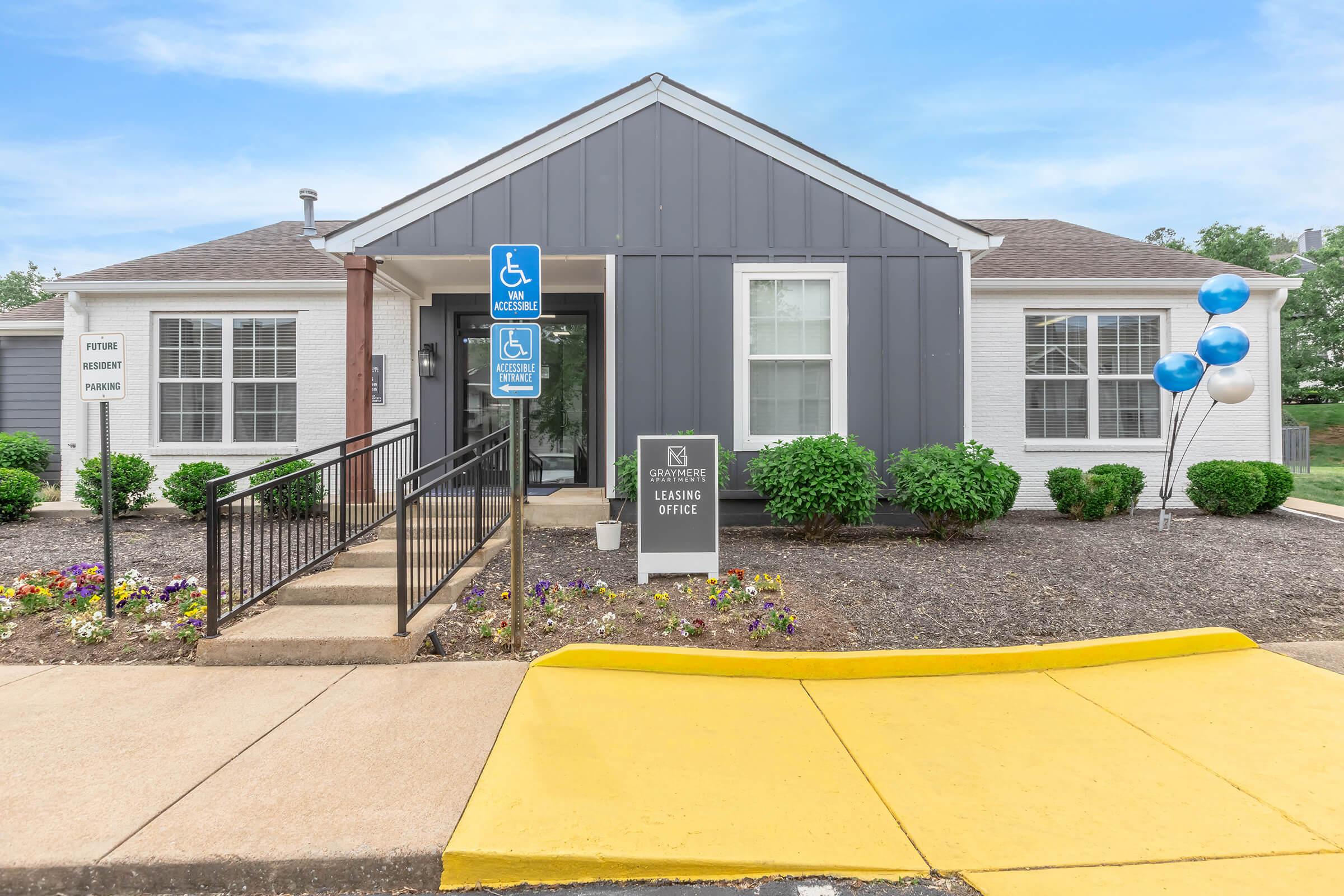 A leasing office building with a gray and white exterior, accessible ramp, and landscaped flower beds. The office sign is prominently displayed, and blue balloons are tied to a railing. The area is well-maintained, with clear skies in the background.