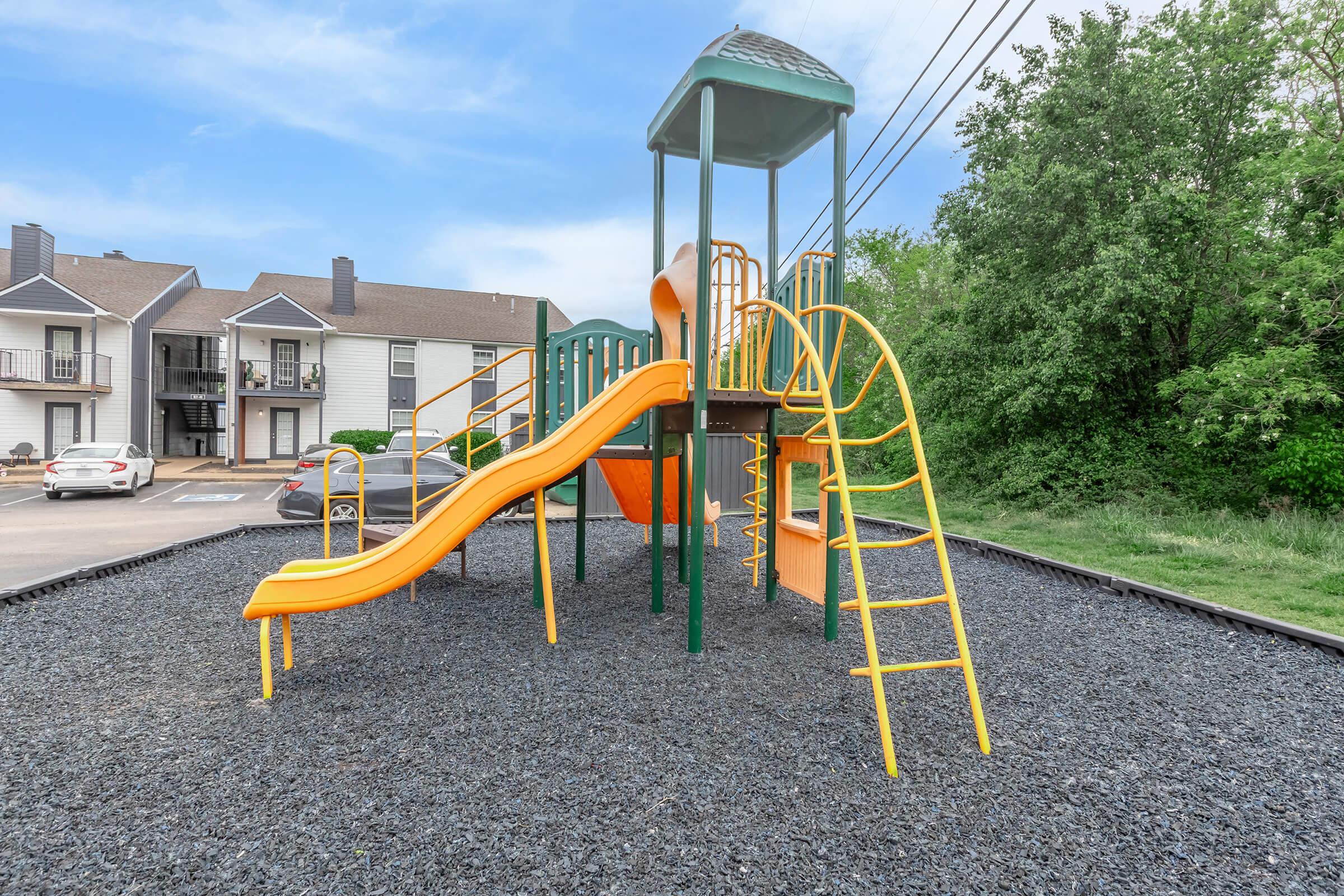 A colorful playground set featuring a green and orange slide, climbing frames, and a canopy. The playground is located on a gravel surface near a residential area, with buildings visible in the background and greenery surrounding the site.