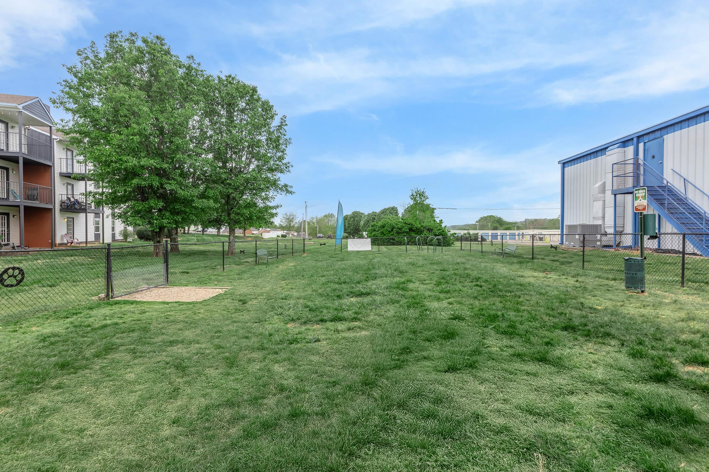 A spacious green yard area surrounded by a fence, featuring a small white monument or structure in the center. Near the yard, there are residential buildings on one side and a blue industrial building on the other. The sky above is clear with a few clouds.
