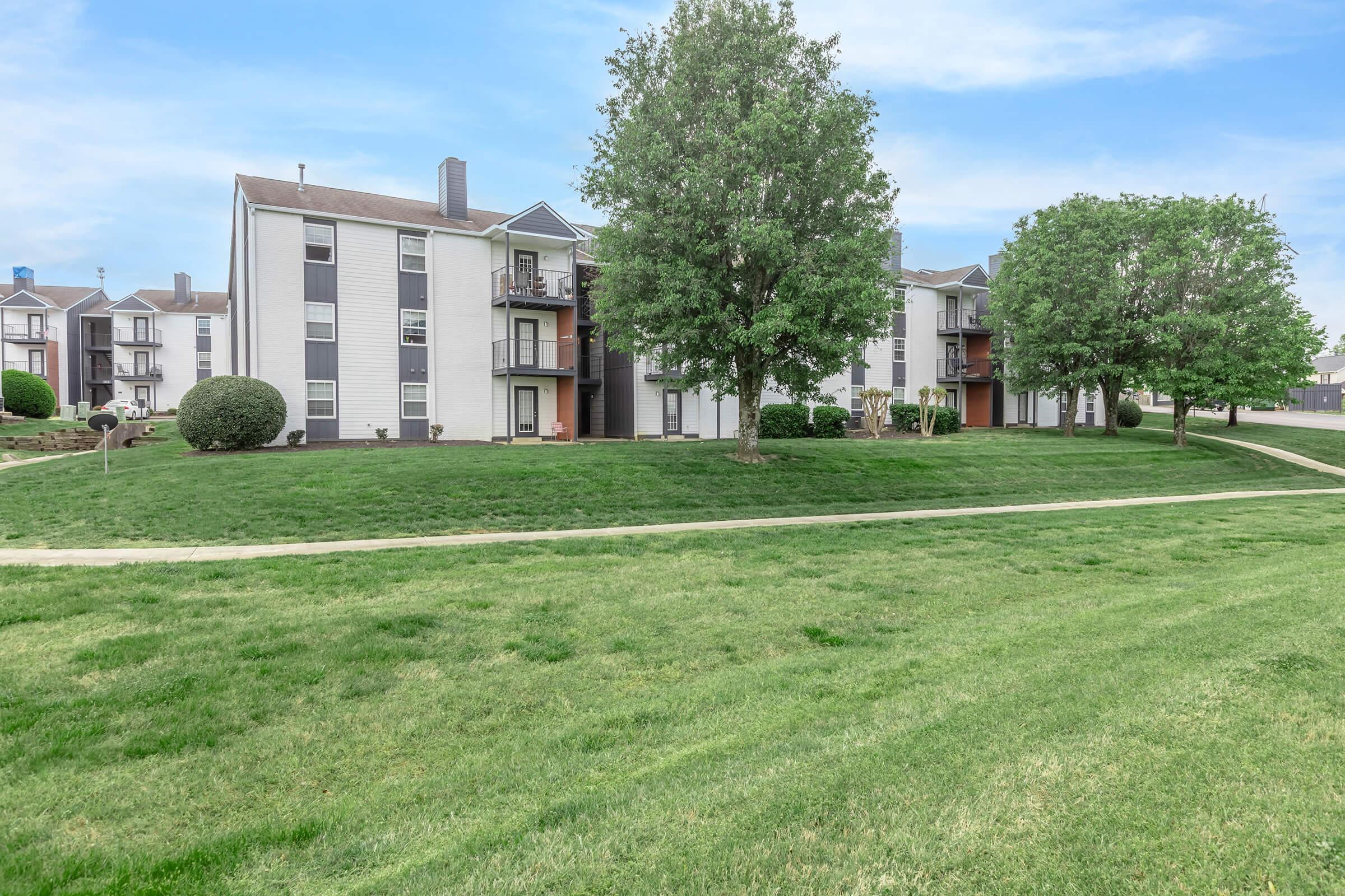 A grassy area in front of several multi-story residential buildings. The buildings are light-colored with balconies, surrounded by neatly trimmed trees and shrubs. A walking path runs across the lawn, providing access to the buildings. The sky is clear with light clouds.