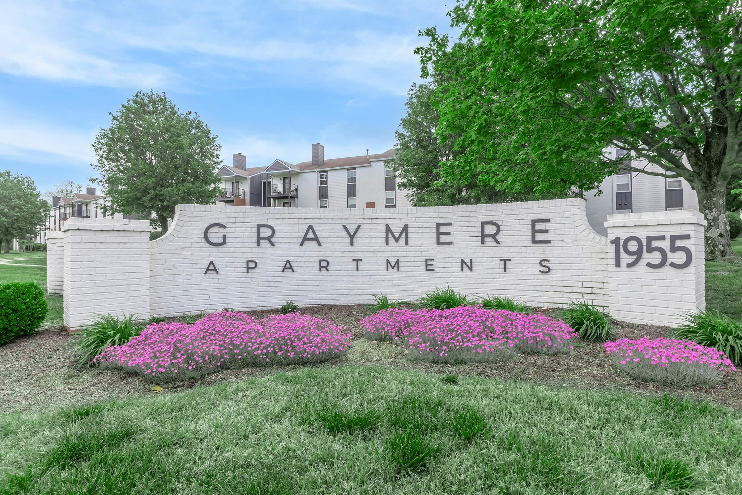 Sign displaying "Graymere Apartments" along with the address "1955", surrounded by green landscaping and colorful flower beds. The background shows residential buildings, contributing to a welcoming atmosphere.
