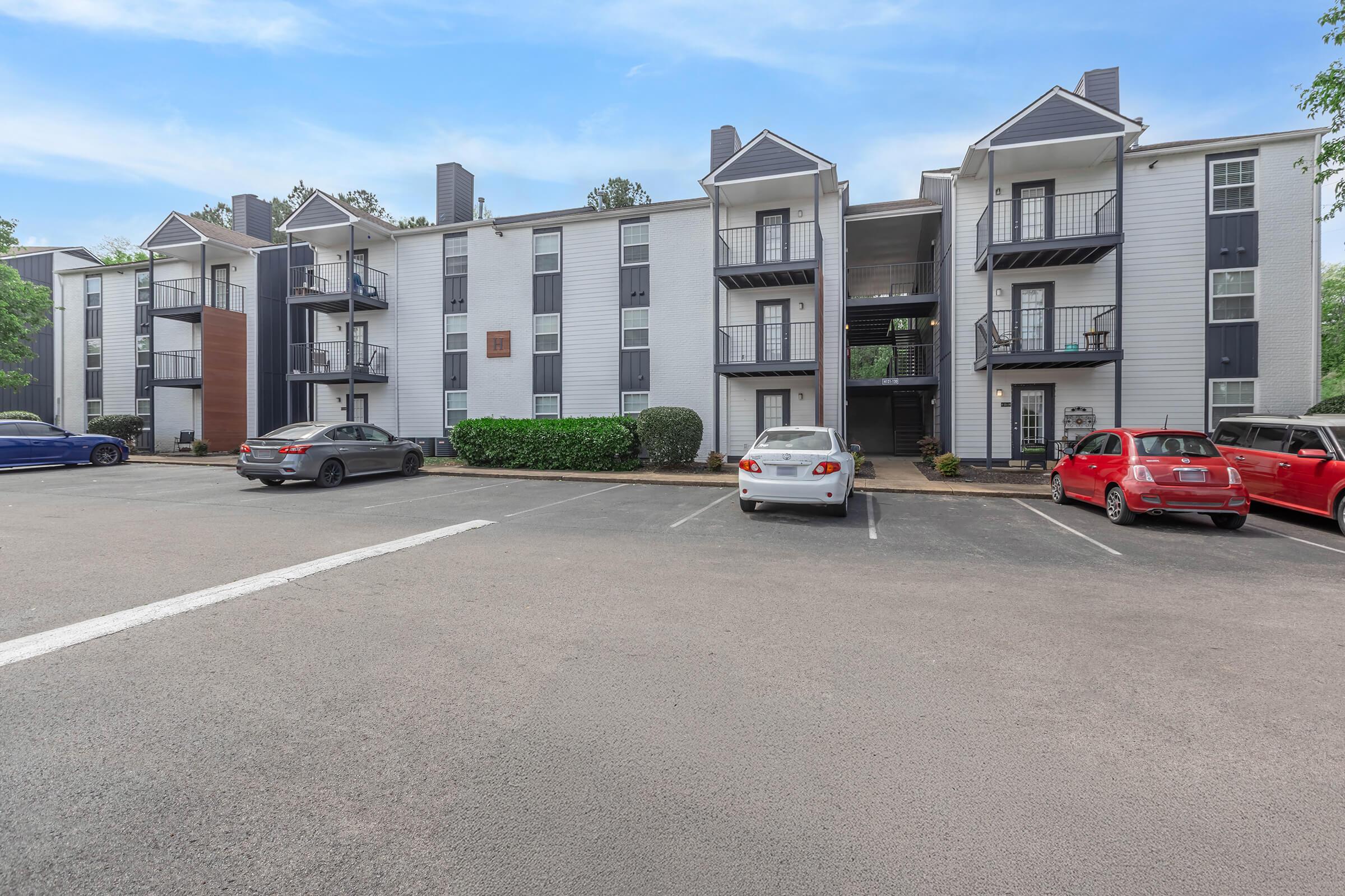 Four-story apartment building with a mix of grey and white siding. The building features balconies and is surrounded by green shrubbery. A parking lot in front has several parked cars, including a blue sedan, a white car, and a red car. Trees and a clear blue sky are visible in the background.