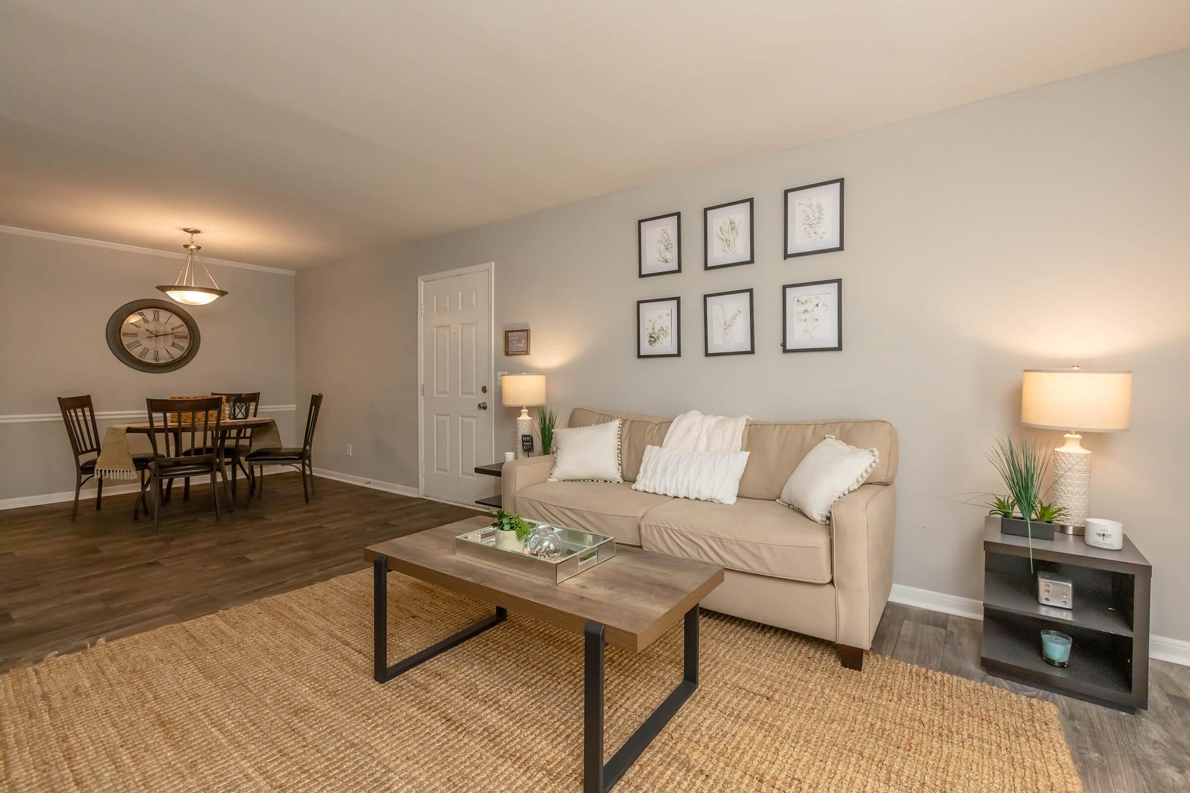 A cozy living room featuring a light beige sofa with decorative pillows, a wooden coffee table, and a woven area rug. In the background, there's a dining area with a round clock on the wall and a wooden dining table with chairs. Soft lighting from table lamps enhances the inviting atmosphere.