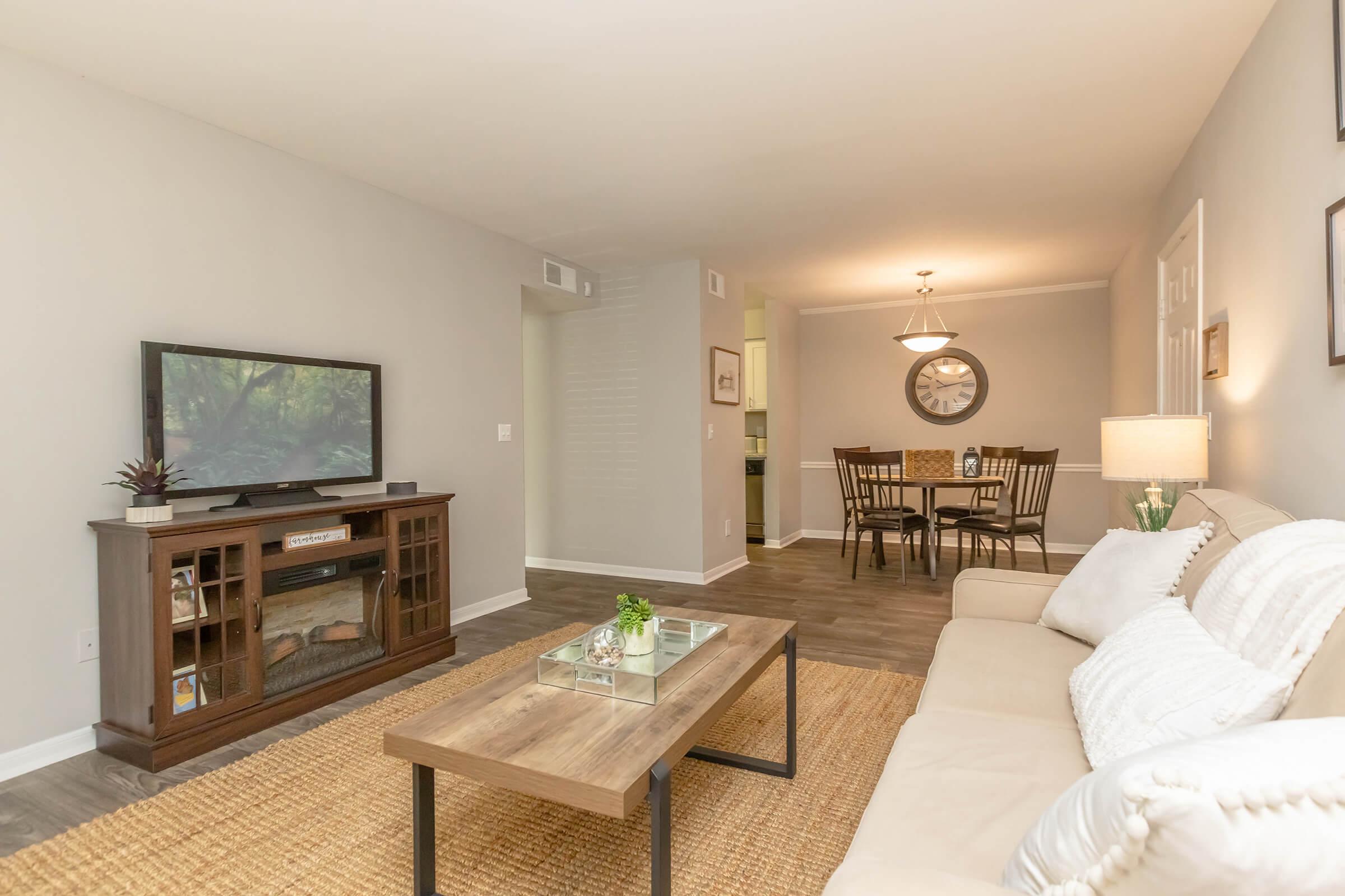 A cozy living room featuring a beige sofa, a wooden coffee table, and a television on a dark wood unit. In the background, there's a dining area with a round table and chairs, illuminated by a chandelier. Neutral-colored walls and a jute rug add warmth to the space.