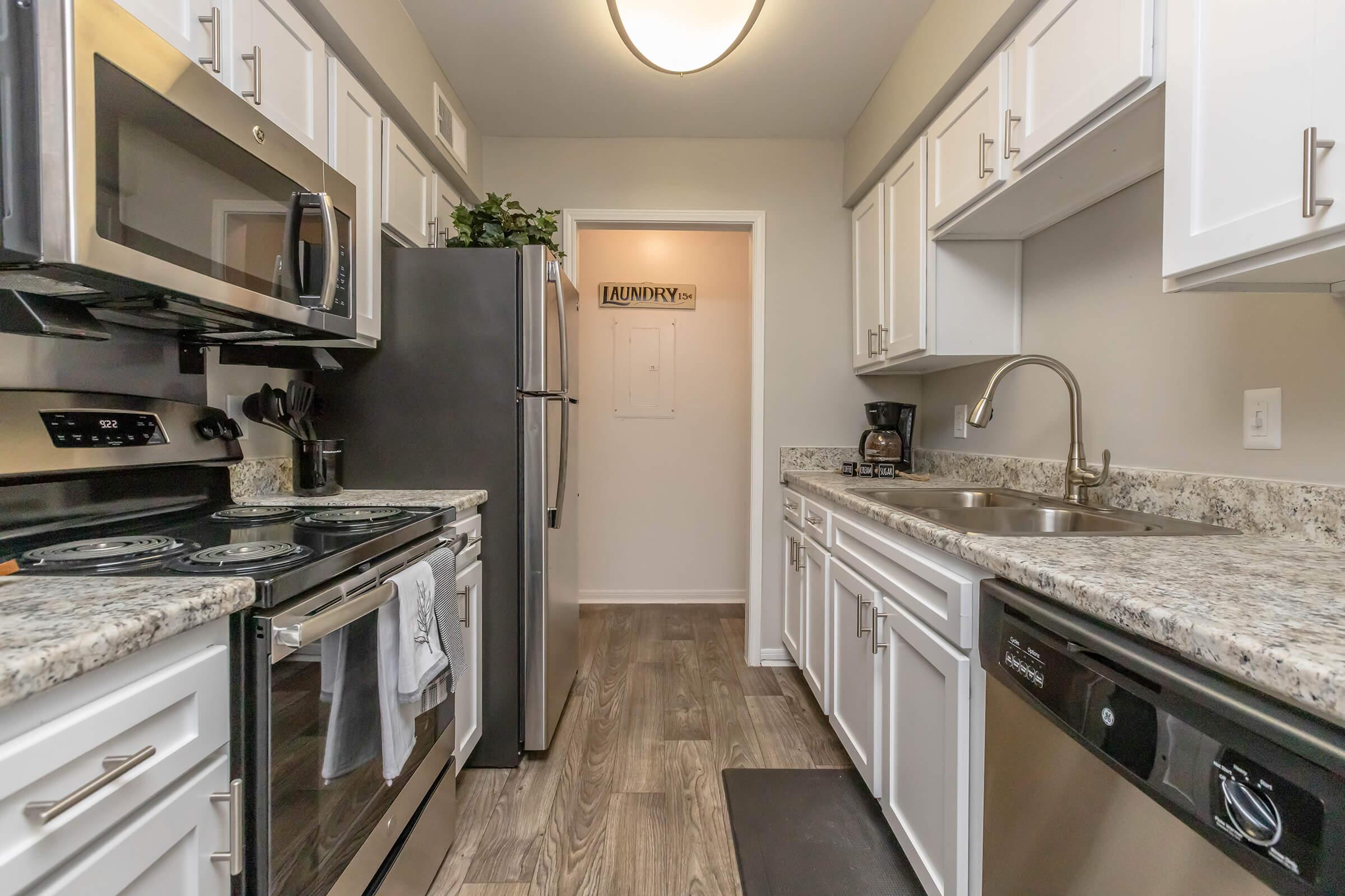 A modern kitchen featuring white cabinets, granite countertops, stainless steel appliances including a microwave, oven, and dishwasher. The space has a sink with a chrome faucet and a coffee maker on the countertop. A door labeled "LAUNDRY" leads to a laundry area in the background.