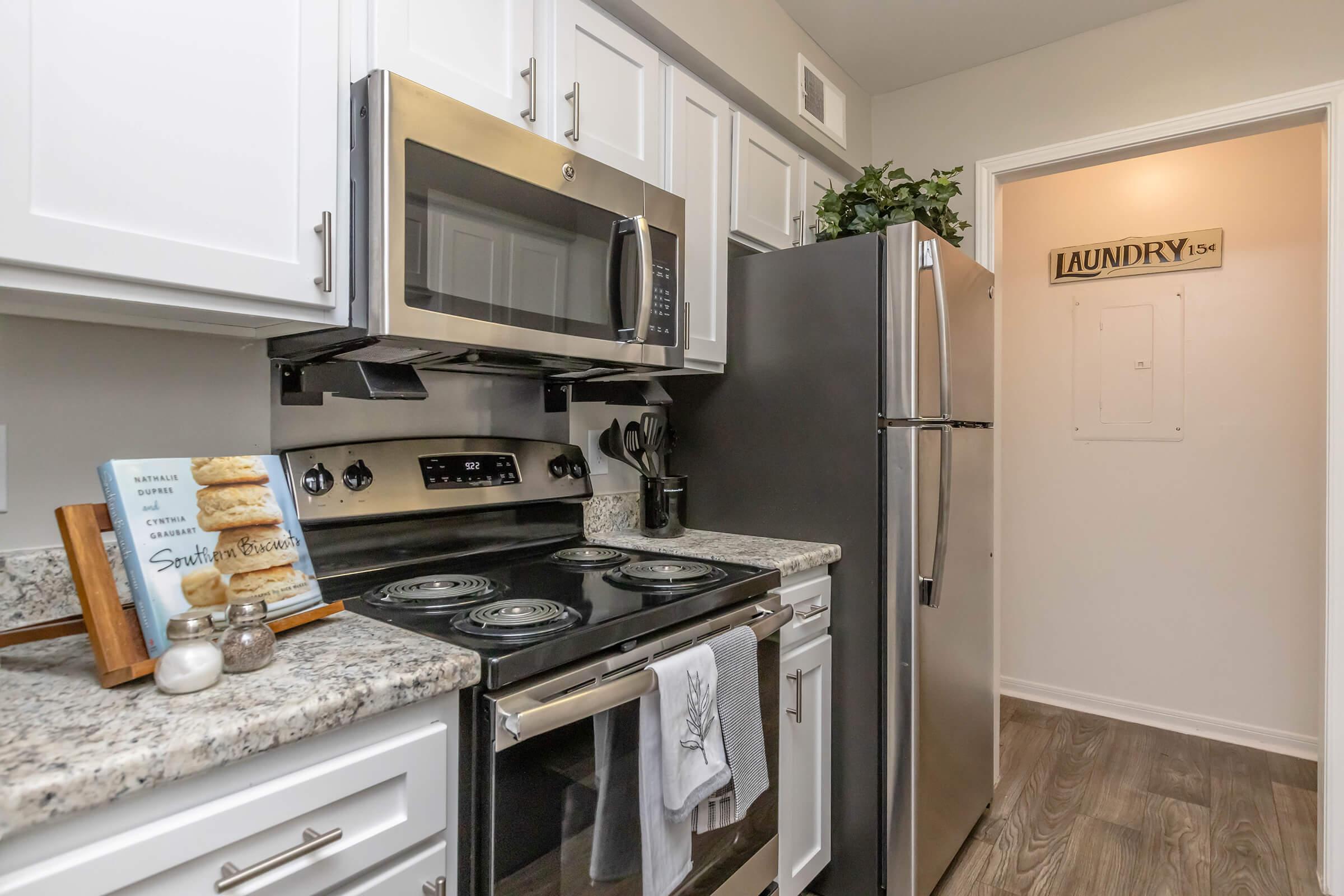 Modern kitchen with white cabinets, stainless steel appliances, and granite countertops. Features a microwave, oven, and a refrigerator. A cookbook sits on the counter next to decorative items. An adjacent laundry room door is visible, labeled "LAUNDRY." Soft lighting enhances the inviting atmosphere.