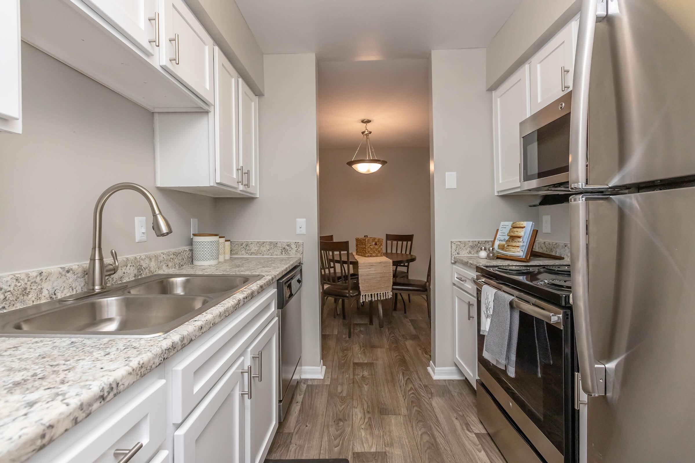 A modern kitchen featuring white cabinetry, stainless steel appliances, and a granite countertop. The sink is placed under a window, and a dining area with wooden chairs and a table is visible in the background, illuminated by a hanging light fixture. The floor is made of wood-like tiles.