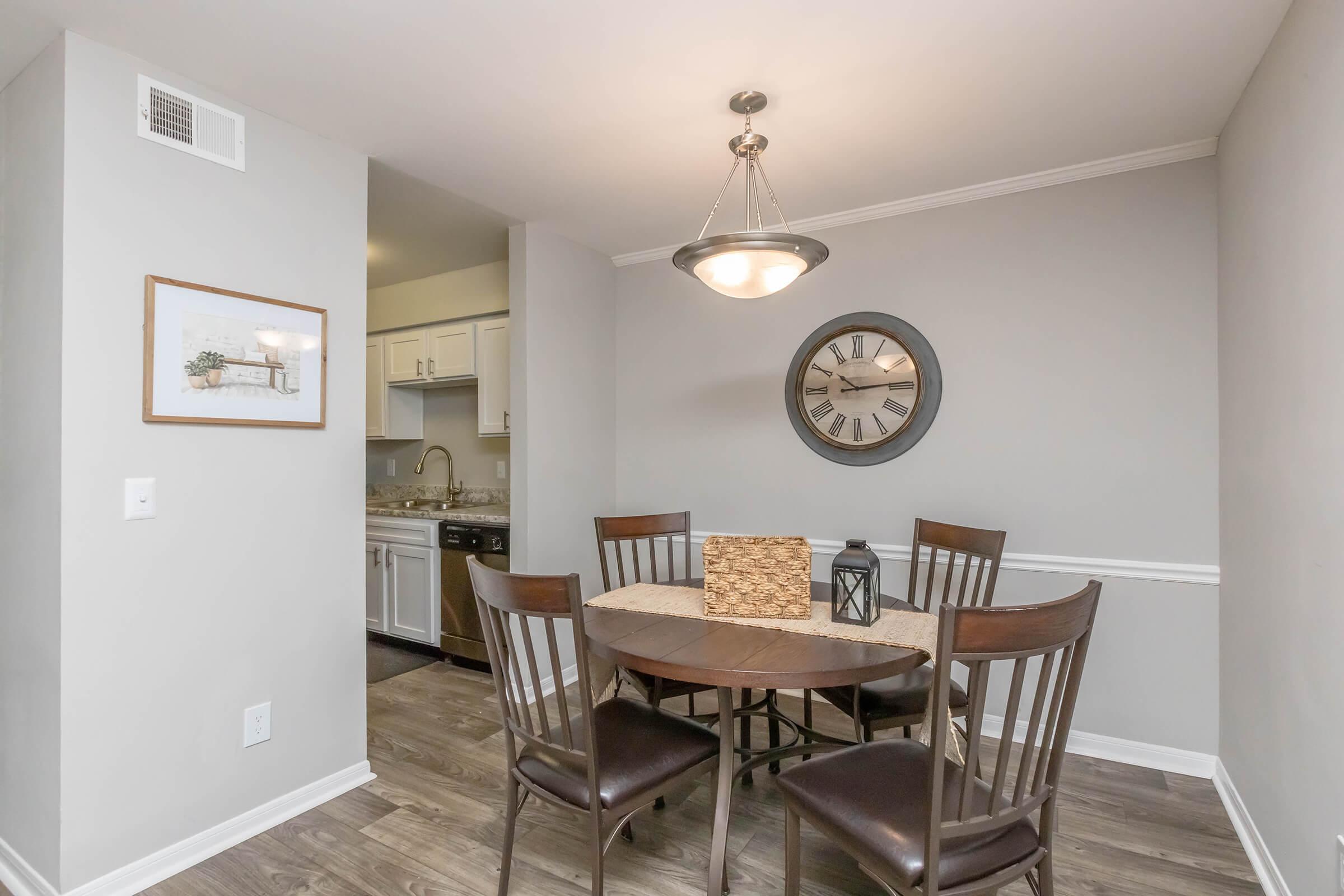 Cozy dining area featuring a round table surrounded by four chairs, a decorative centerpiece, and a lantern. The walls are painted in soft gray, with a clock and artwork adding charm. A kitchen is visible in the background, enhancing the inviting atmosphere of the space.
