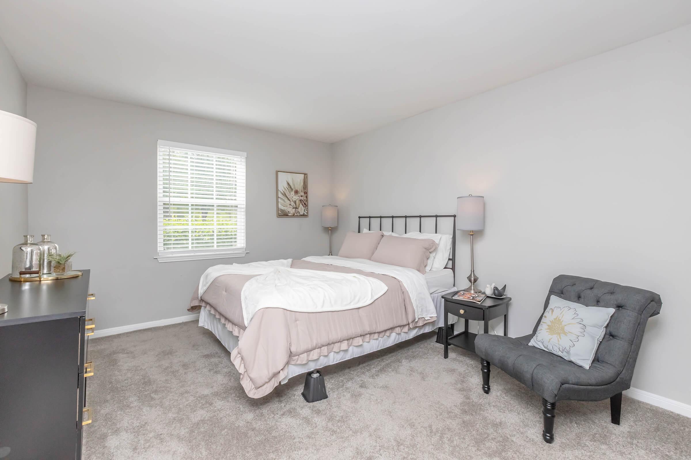 Cozy bedroom featuring a bed with light bedding, two bedside lamps, and a small armchair. A window allows natural light, and the walls are painted in soft gray. A dresser is positioned against one wall, adding to the room's inviting atmosphere.